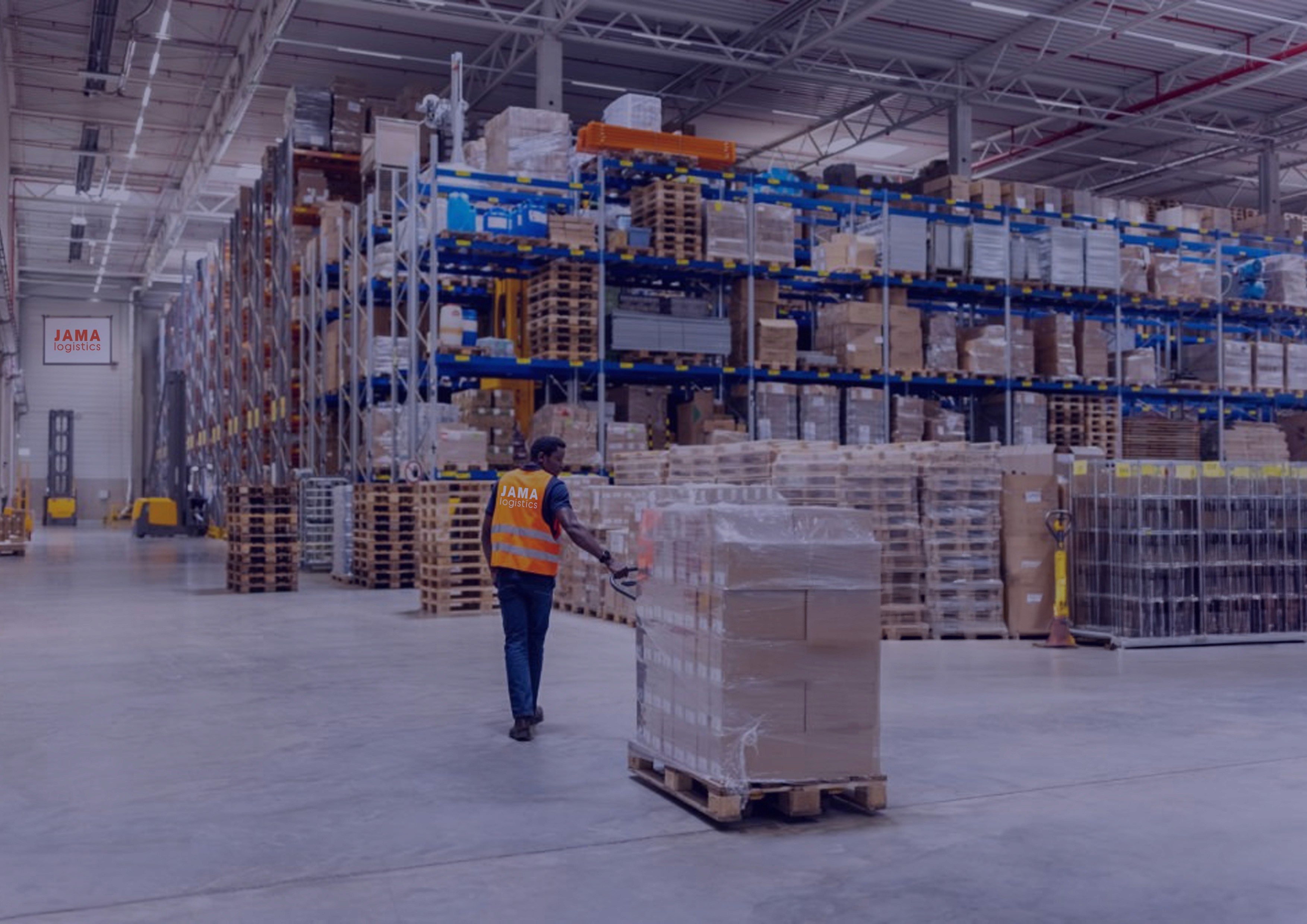 Image of a man wheeling a pallet load through a warehouse with jama logistics logo on the wall