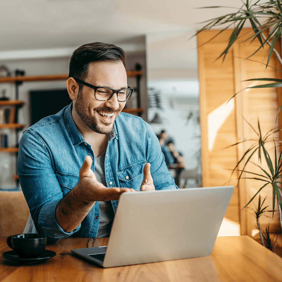 A man in a casual denim shirt smiling and gesturing towards his laptop screen, indicating a virtual meeting or online collaboration.