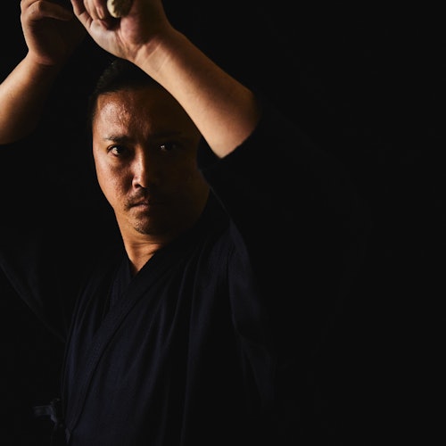 Een persoon in een donkere outfit houdt een zwaard boven zijn hoofd tegen een zwarte achtergrond, met een geconcentreerde uitdrukking.