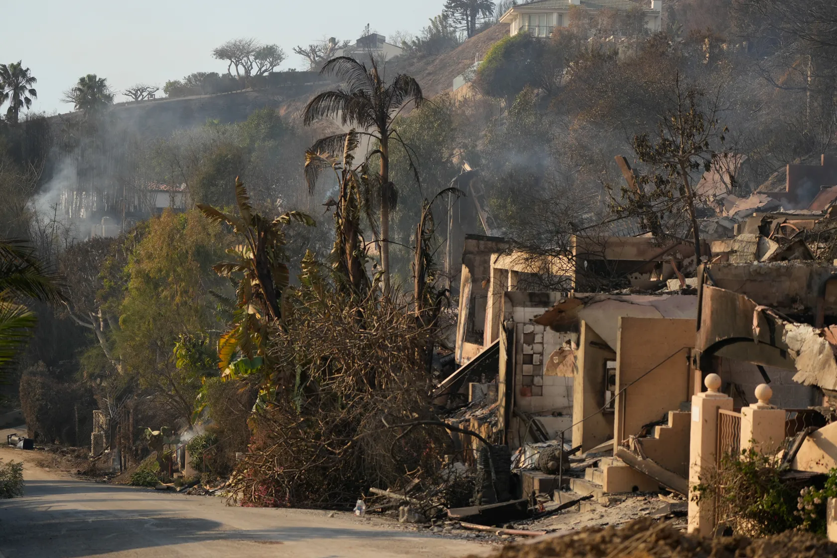 A neighborhood in Malibu shows the charred remains of a custom home, with smoldering debris and surrounding scorched vegetation from the Pacific Palisades fires.