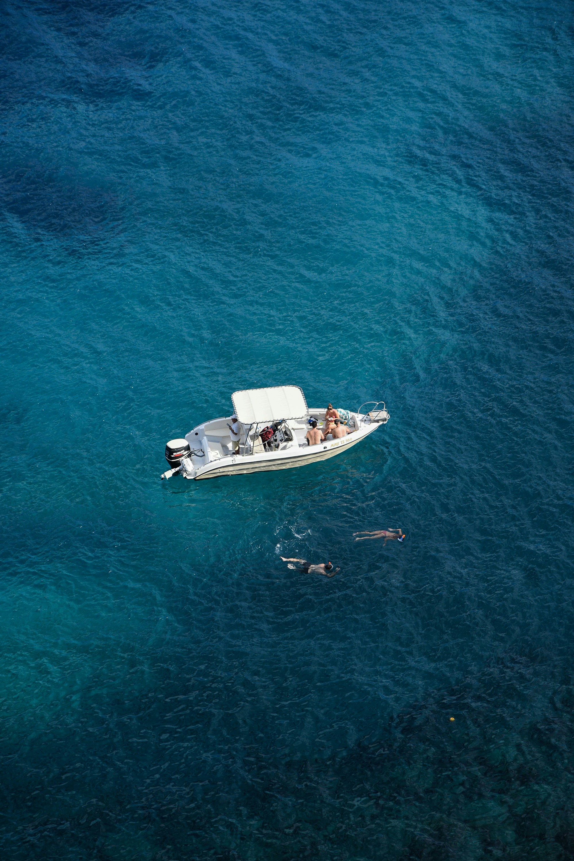 A white motorboat floats on crystal-clear blue waters as two people swim nearby, capturing the essence of a yachting experience and island retreat.