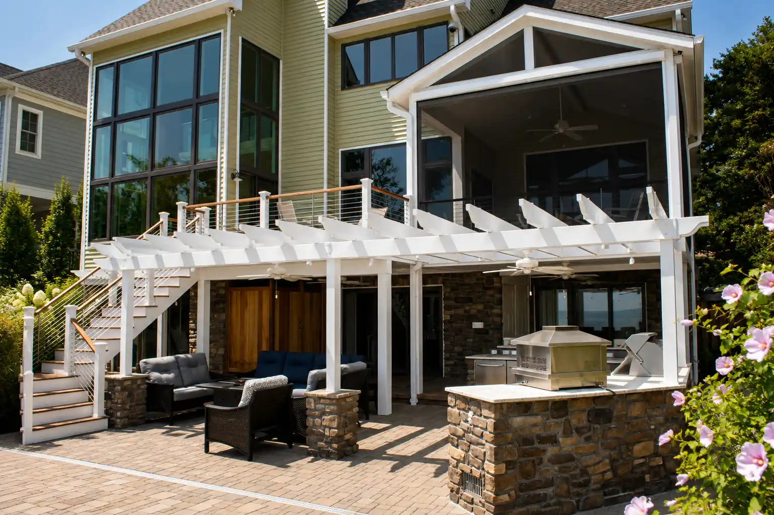 Rear exterior featuring an elevated deck, pergola-covered patio, outdoor kitchen, seating area, and landscaped poolside space.