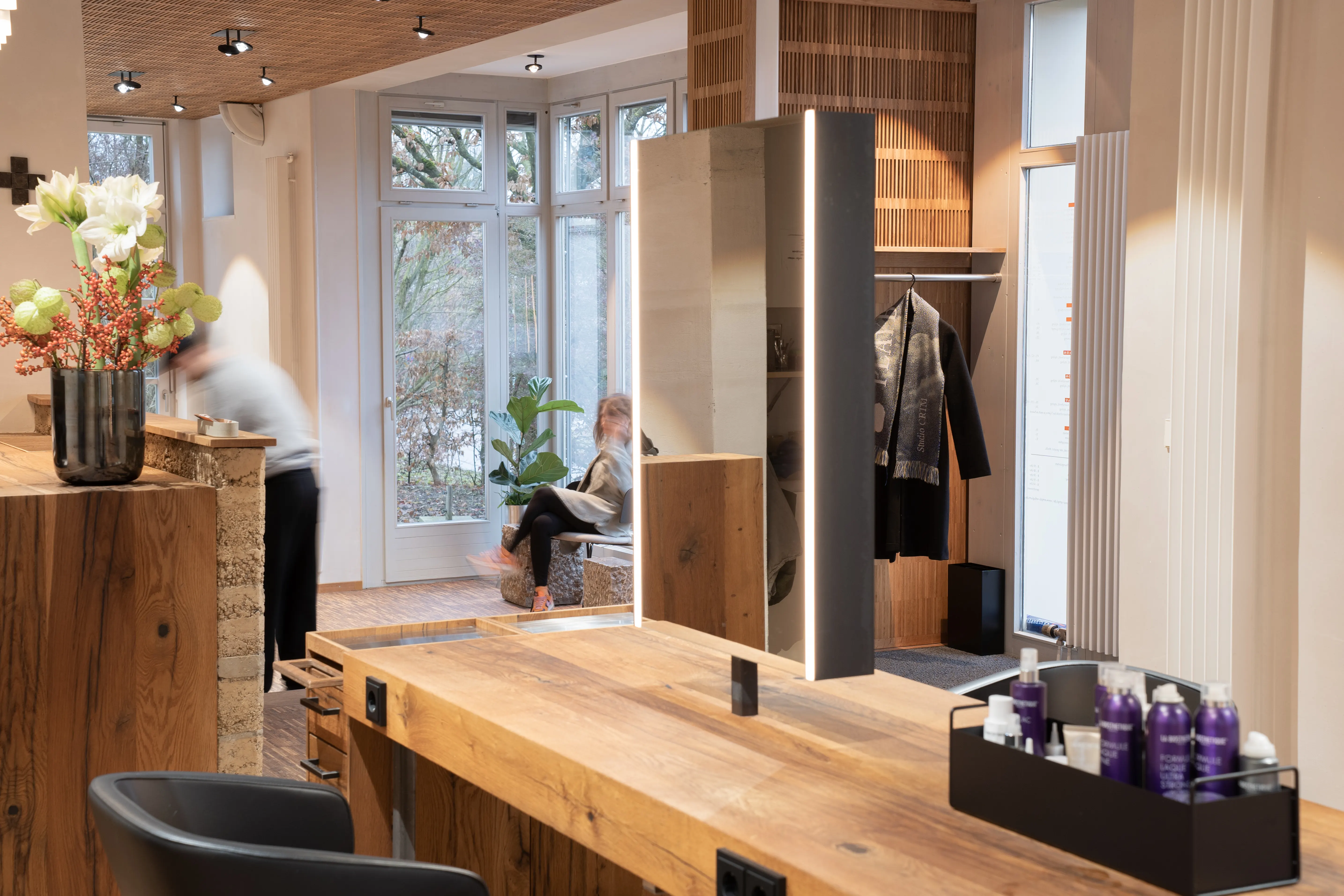 Interior of EMPL FRISEURE with an oak workbench, double-sided illuminated mirror, hair products in a black tray and a window seating nook with a reading customer.