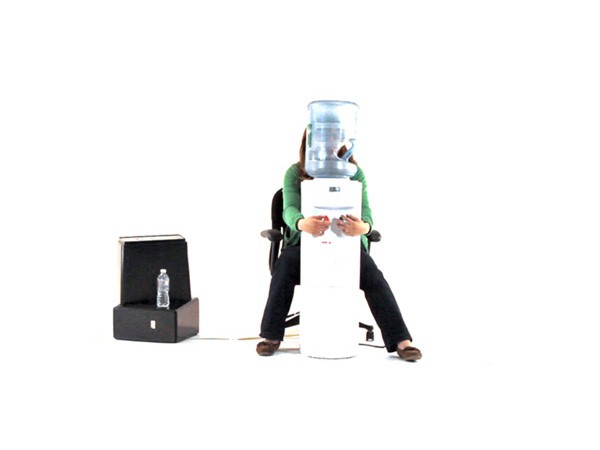 A person sits in a black office chair behind a white water cooler with a transparent plastic container on top. The person is wearing a green long-sleeved shirt and dark pants. A black box with a water bottle on top sits to the left of the cooler. The background is plain white.