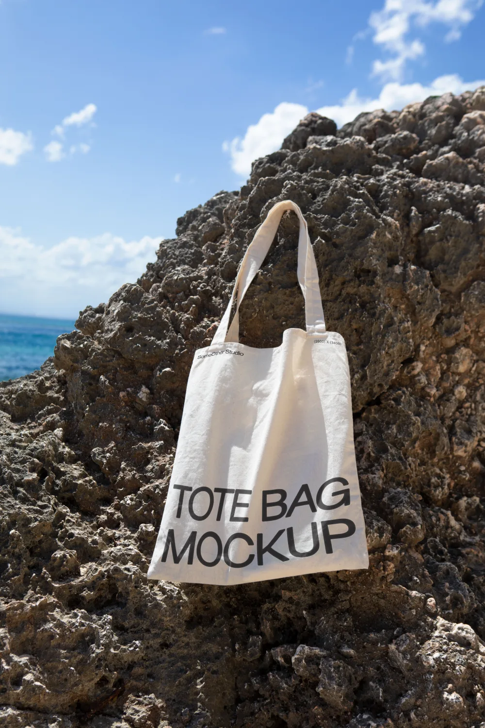 A natural cotton tote bag mockup hanging on a textured rock surface, set against a bright sunny backdrop with a clear blue sky and ocean waves in the distance.