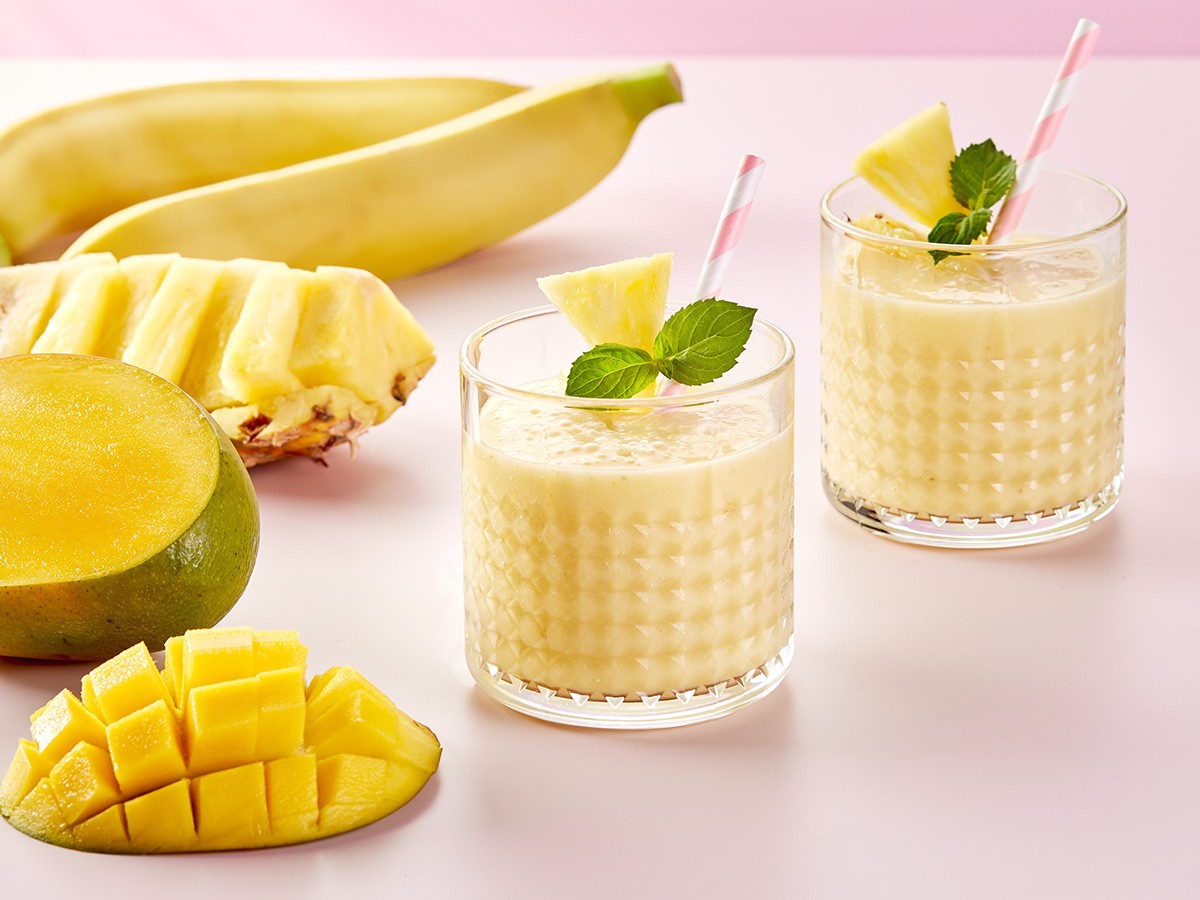Two glasses of creamy mango banana smoothie garnished with pineapple slices and mint leaves sit on a pale pink surface, surrounded by fresh mango, banana, and pineapple pieces.