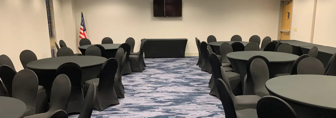 A conference room with round tables and black chairs arranged for a meeting or event, with a blue patterned carpet and an American flag in the background.