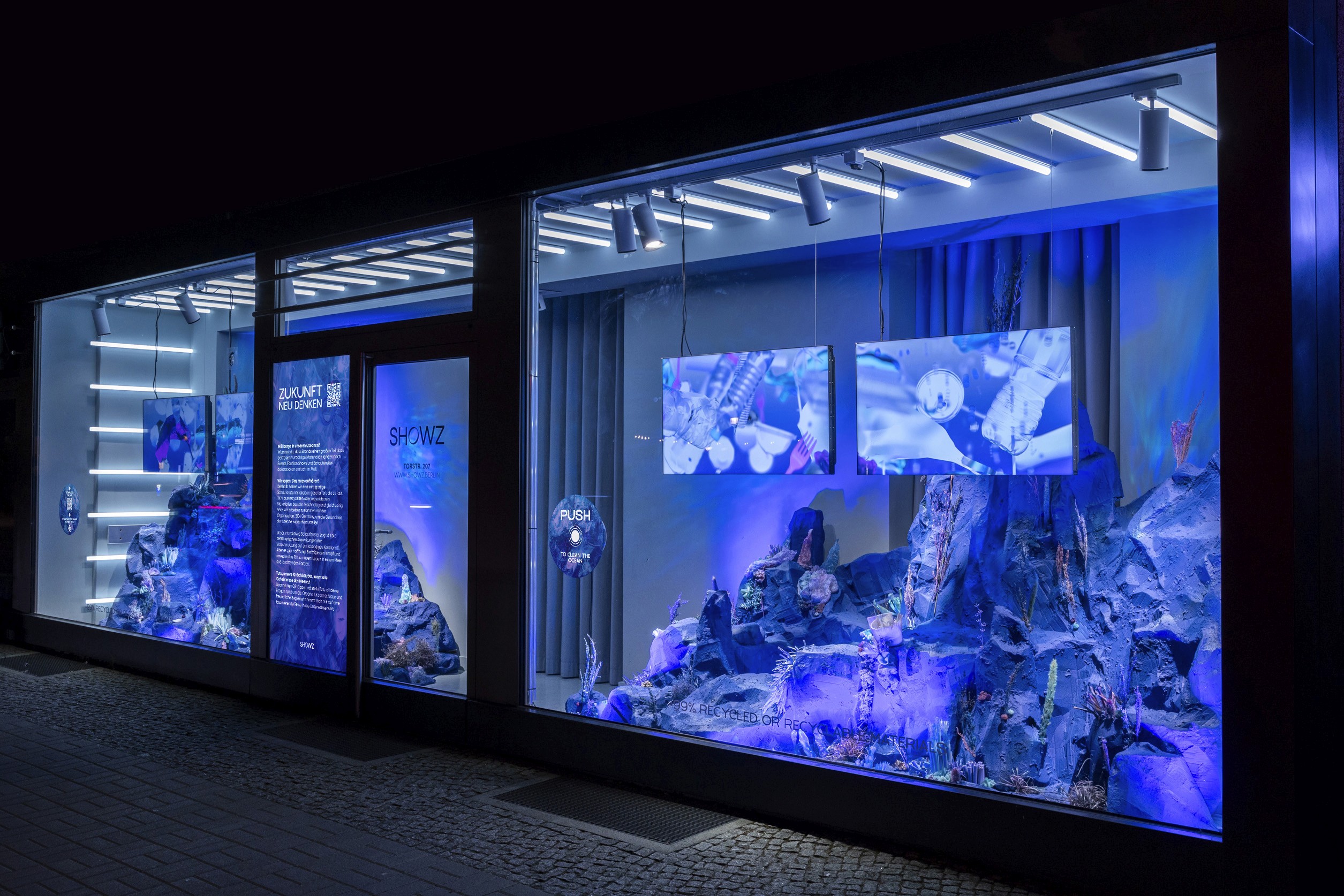 Futuristic retail window display at night for SHOWZ event, featuring immersive blue lighting, recycled material sculptures resembling underwater landscapes, and suspended digital screens showcasing innovation and sustainability themes