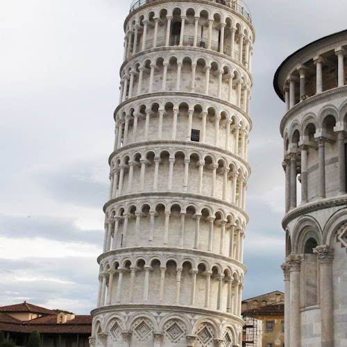 The Leaning Tower of Pisa next to the edge of the Pisa Cathedral under a cloudy sky.