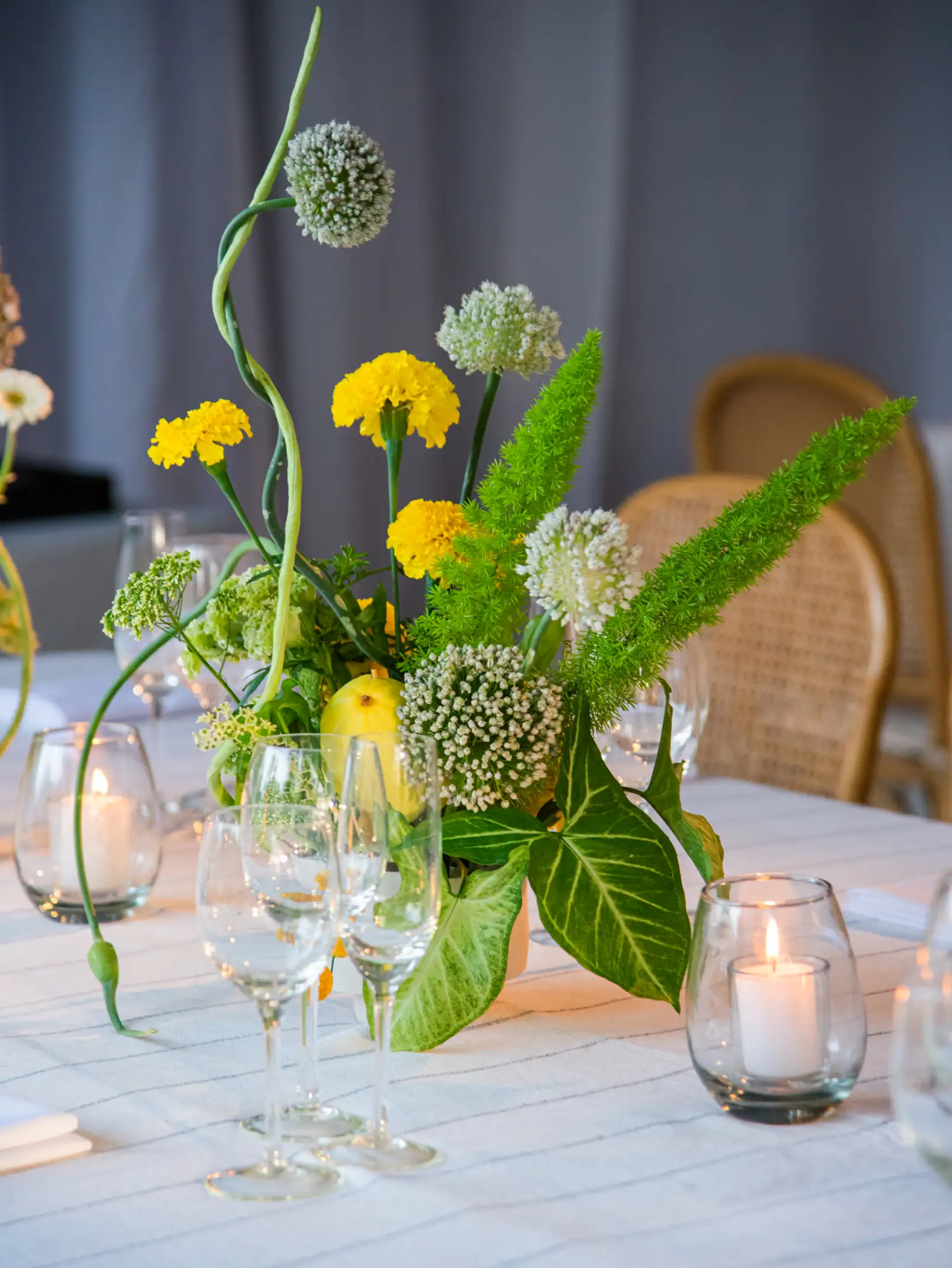 Centro floral con flores amarillas y verdes, limones decorativos y velas sobre mesa vestida en tonos neutros.