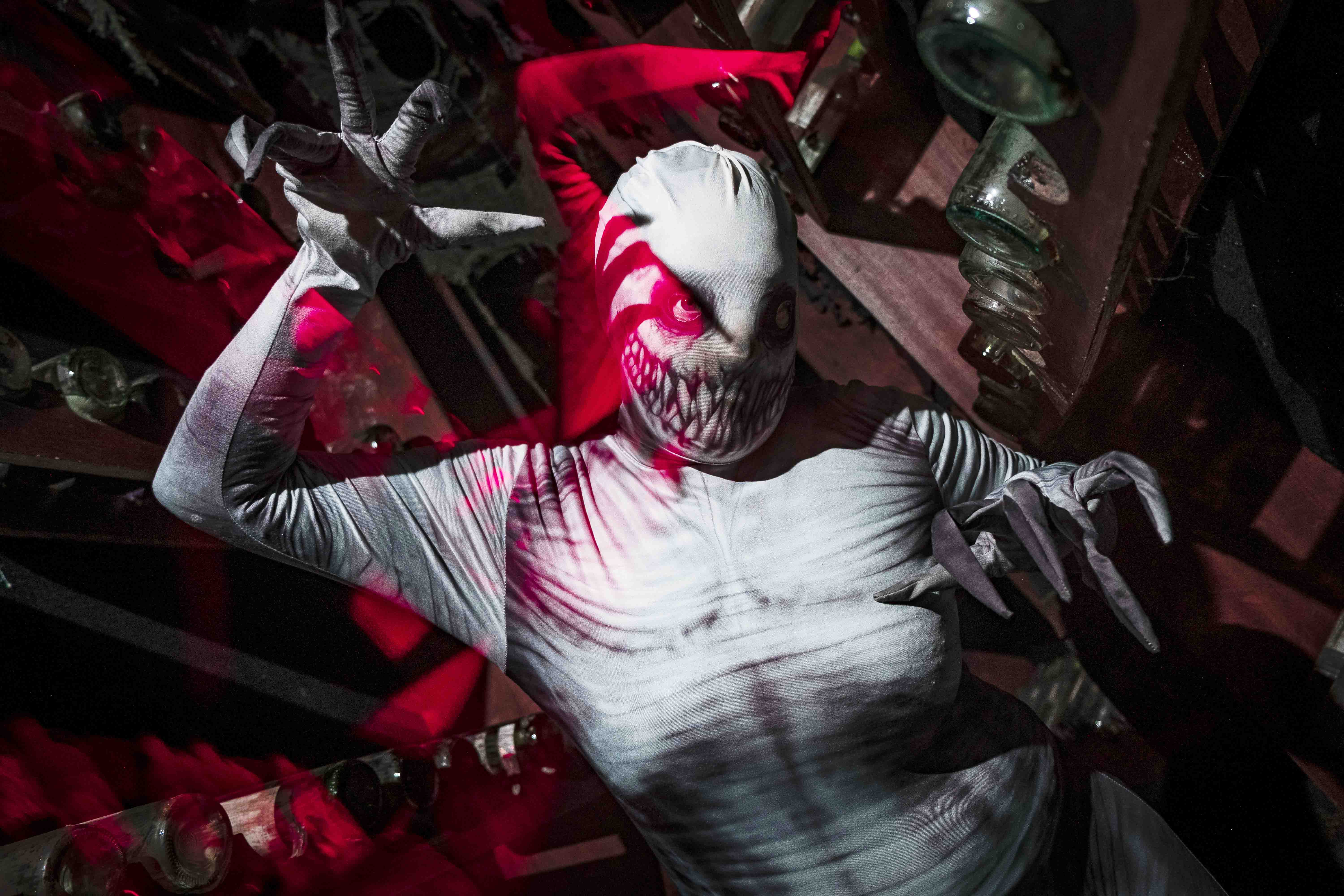 A person in a form-fitting white costume with a menacingly painted face poses against a background of red and dark tones, creating an eerie and dramatic atmosphere.