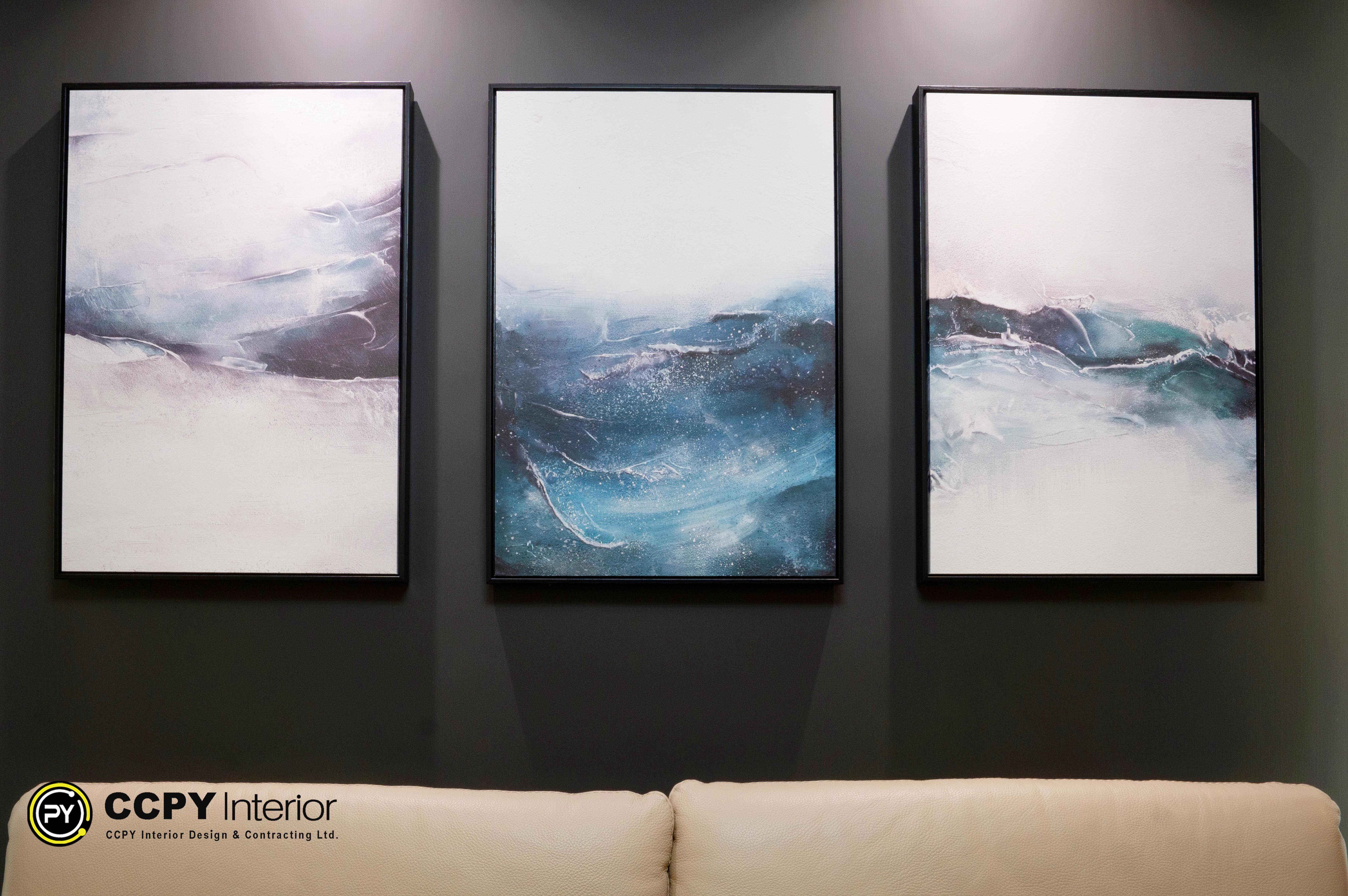The selected region shows a triptych (a set of three panels) of framed wall art featuring an abstract blue and white ocean wave motif mounted on a dark charcoal gray accent wall.Design and Context BreakdownArt Style: Abstract expressionist seascape with fluid, swirling blue, teal, and white tones resembling breaking ocean waves.Framing: Each of the three vertical canvas panels is enclosed in a minimalist, thin black floating frame.Placement: Centered as a focal point on a dark, matte charcoal accent wall directly above a neutral-colored sofa.