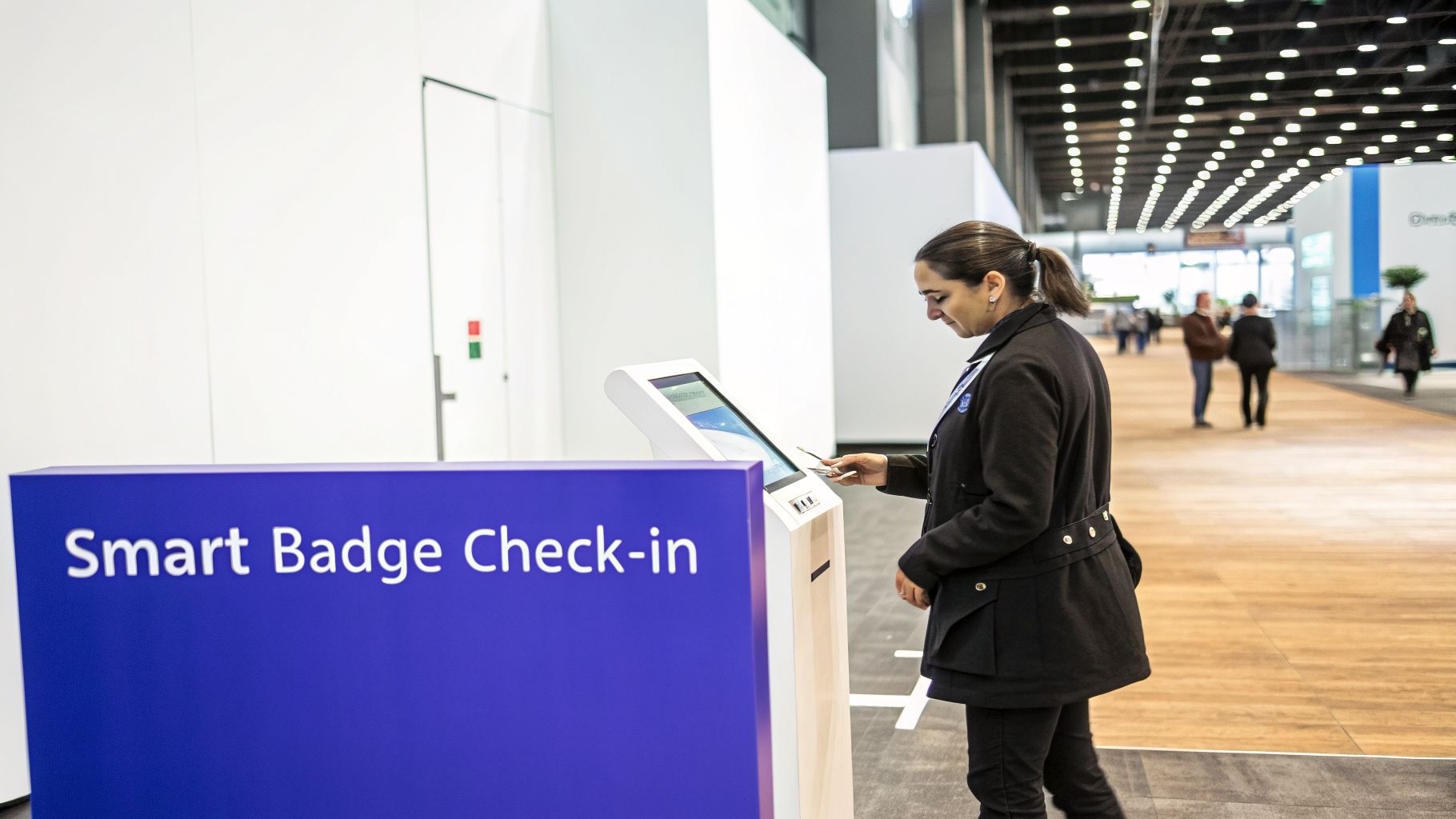 A woman uses a smart badge check-in kiosk at a large event hall with people in the background.