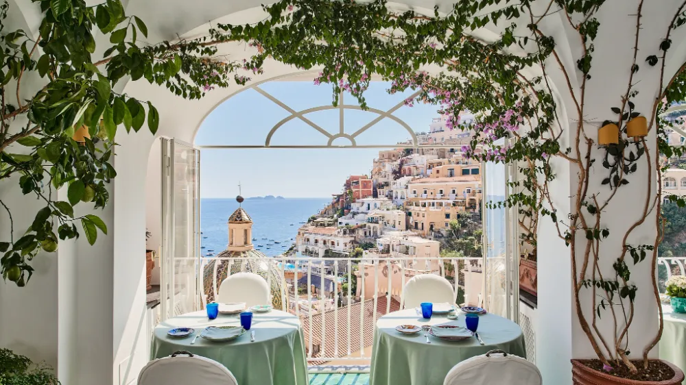 A restaurant terrace with set tables framed by an arch, looking towards Positano's church dome.