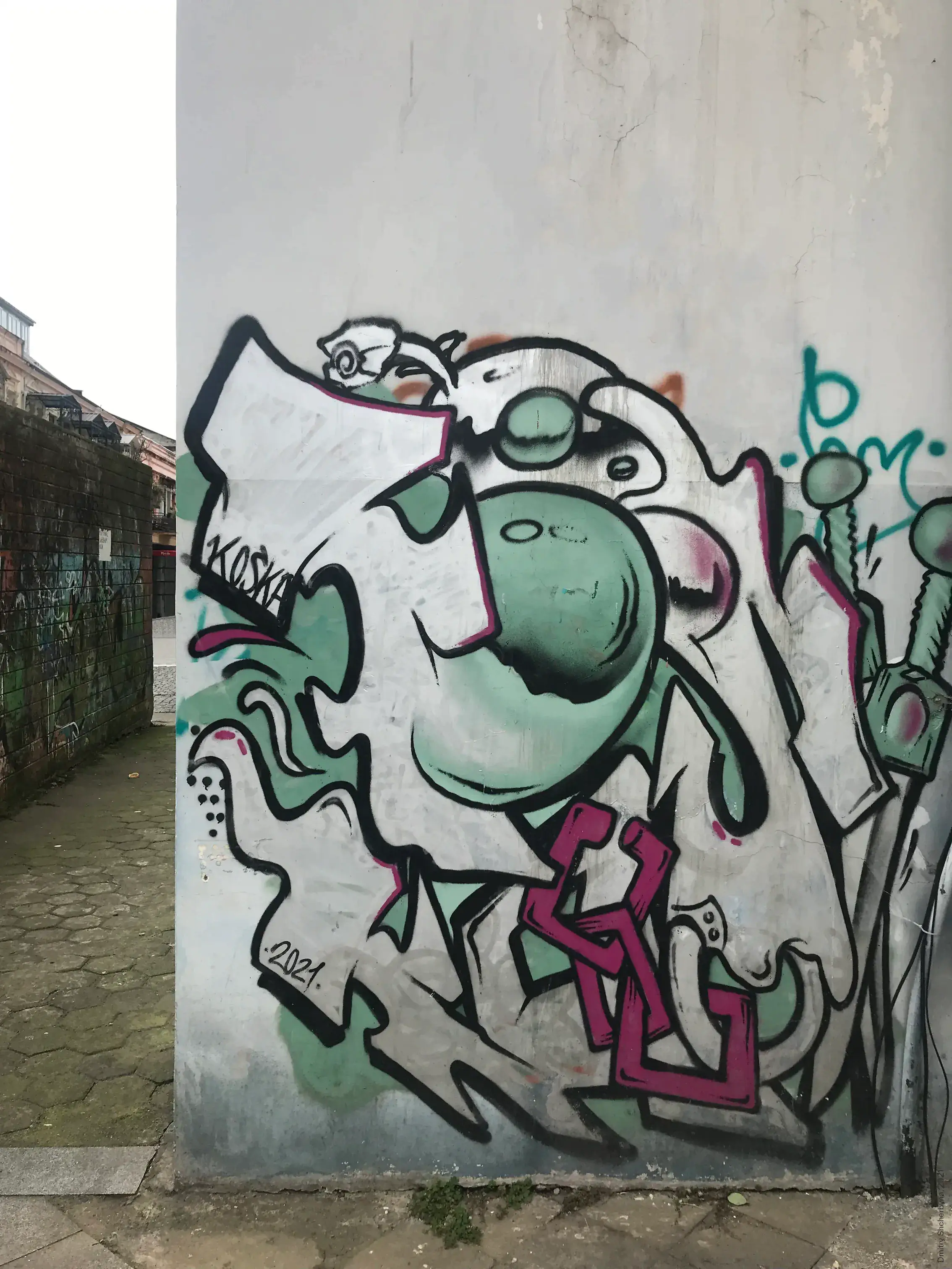 A graffiti of a weathered green wall featuring a round cartoon character with a bulbous nose and wide eyes surrounded by angular letters in mint and pink.