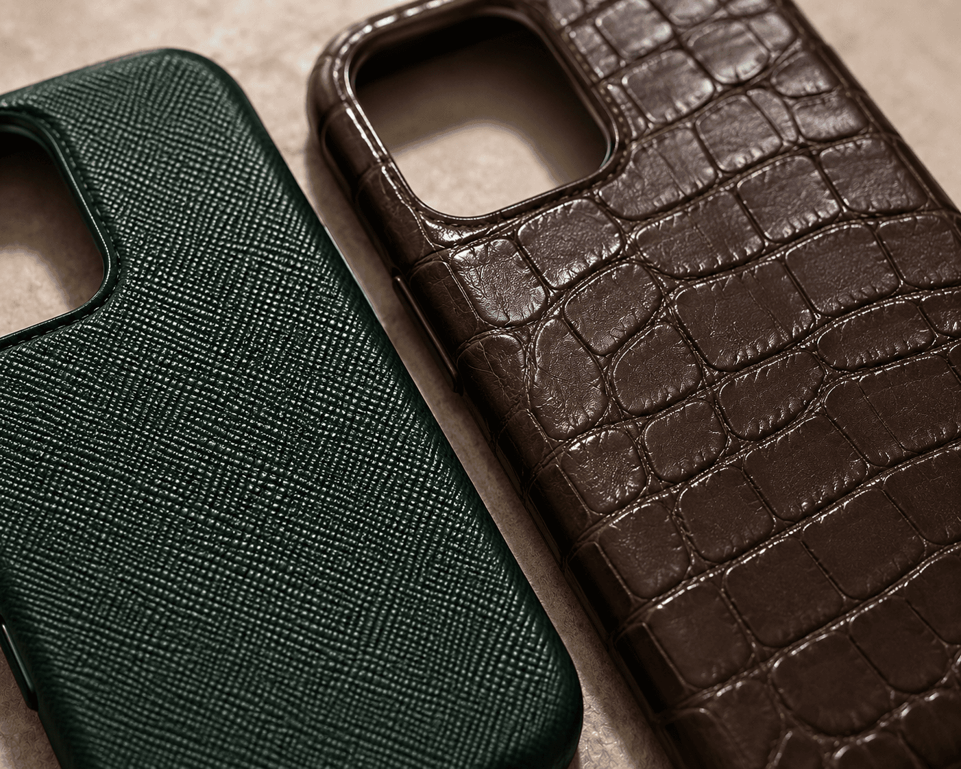 A detailed macro shot focusing on two premium leather smartphone cases lying side-by-side on a light textured surface. On the left, a case features a dark forest green leather with a fine cross-hatch Saffiano texture. On the right, a case highlights a rich, dark brown alligator or crocodile embossed leather with deep, glossy square-scale patterns. The precise camera lens cutouts for both cases are clearly visible at the top.