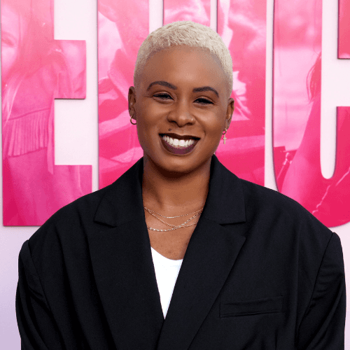 Ashleigh Warren, a person with short, light-colored hair smiles confidently at an event with a pink background.