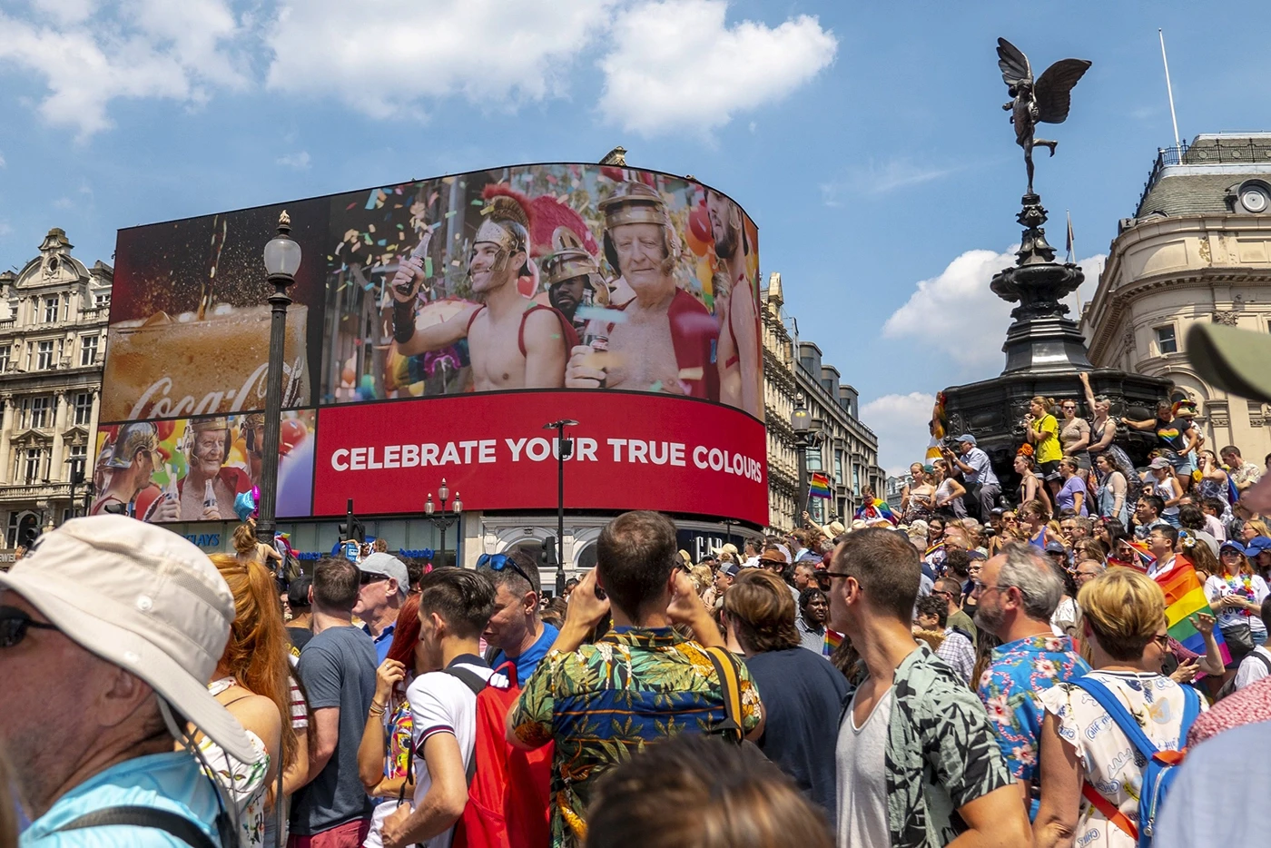 Coca-Cola 'Celebrate Your True Colours' campaign displayed on Piccadilly Circus giant screen during London Pride, large crowd gathered below with rainbow flags visible alongside Eros statue, First Taste advert imagery on LED billboard