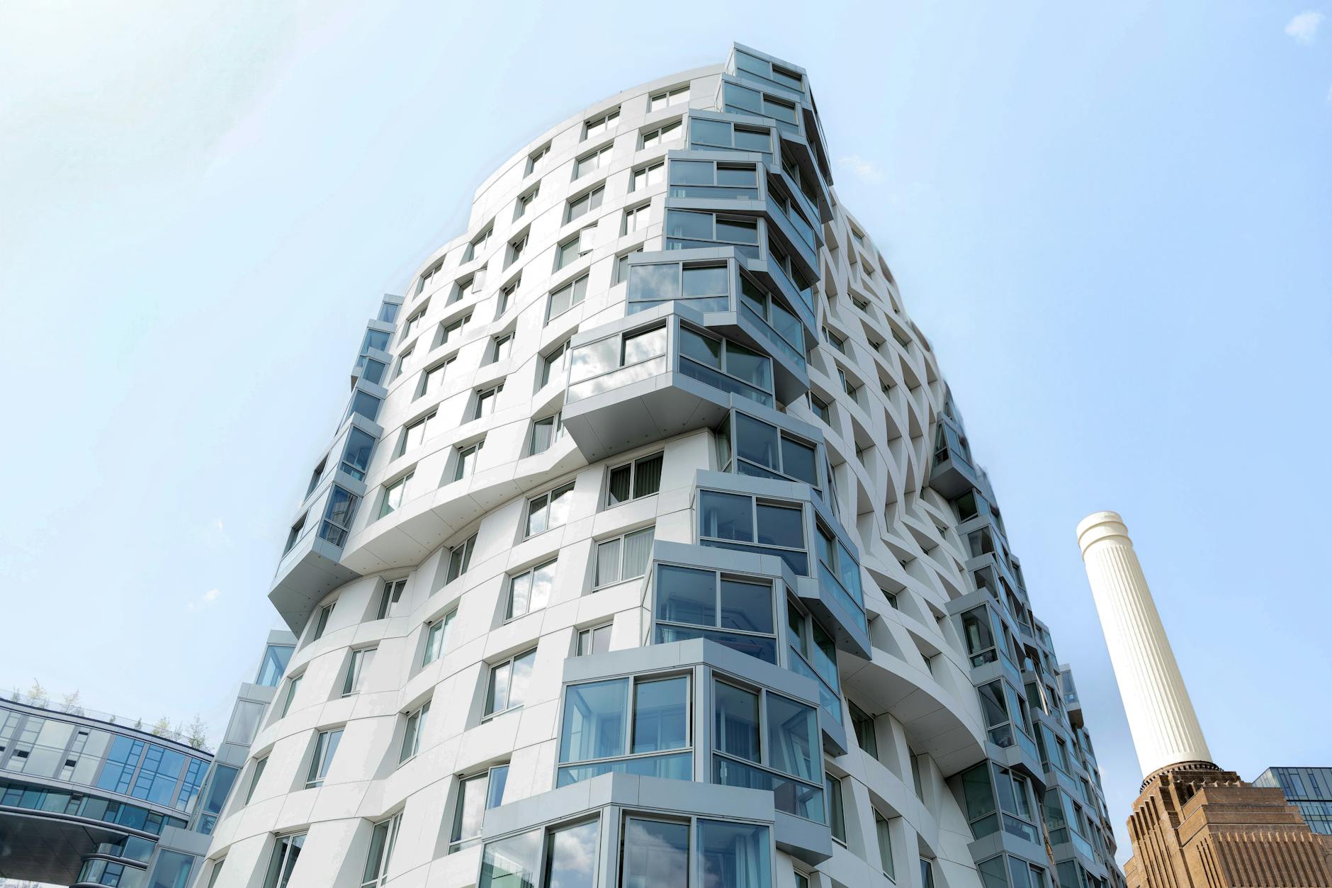 Modern white London apartment block with curved glass façade against blue sky, Battersea chimney behind.