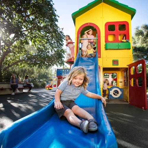 A child slides down a blue slide attached to a multi-colored playhouse, with other children playing on the structure in the background.