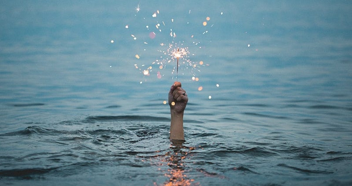 Hand holding a sparkling firework above the surface of a calm blue ocean.