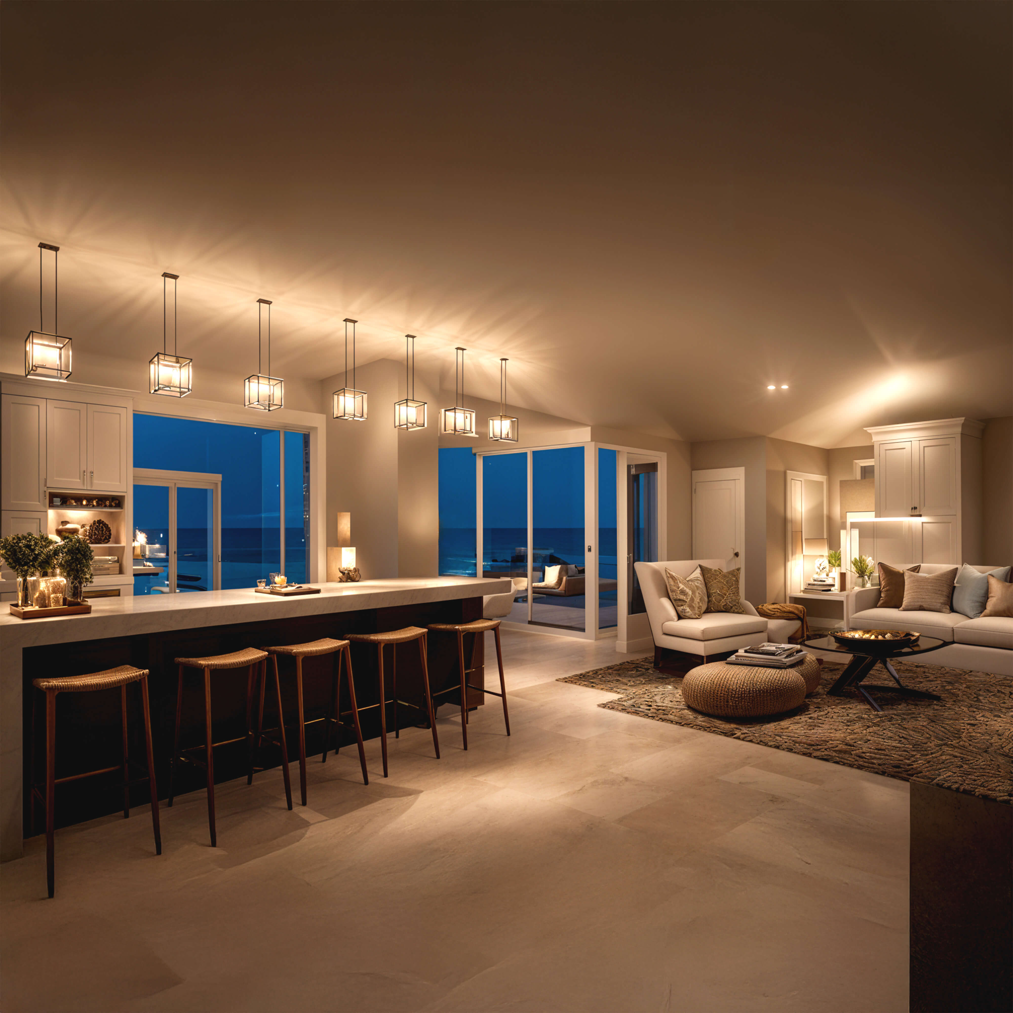 Sophisticated coastal living room and kitchen design with statement pendants, soft neutral furnishings, and expansive glass doors overlooking the ocean at sunset.