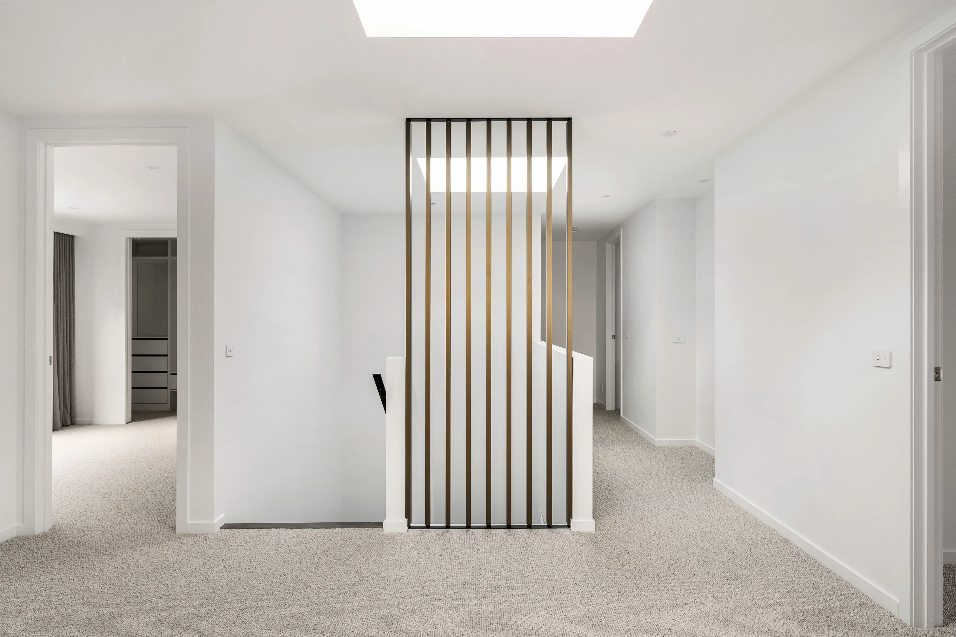 Carpeted upstairs landing with a bronze vertical batten balustrade around the stair void, a skylight overhead and a walk-in robe beyond
