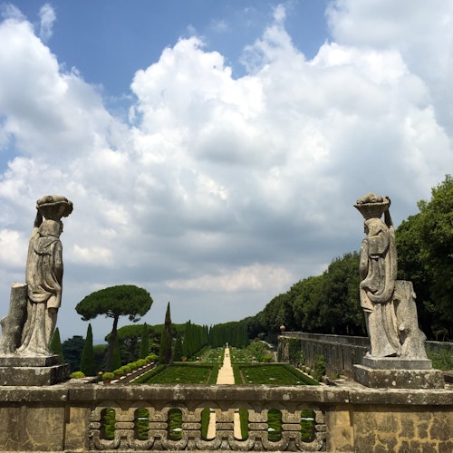 Two stone statues flank a gravel pathway leading into a manicured garden with trimmed hedges, under a partly cloudy sky.