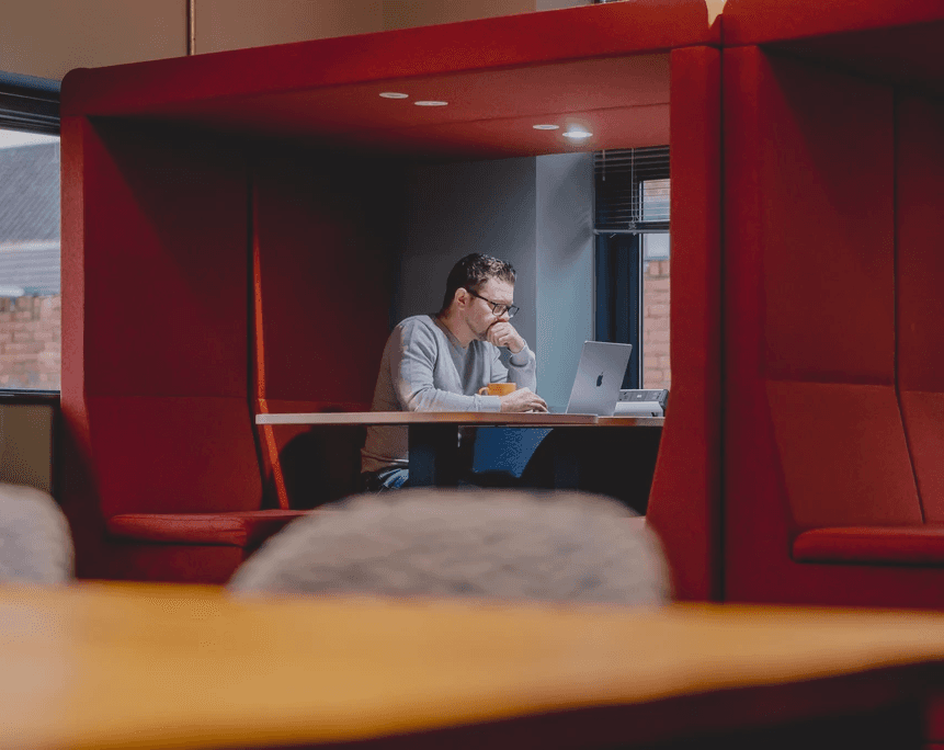 Person seated in a red enclosed office booth, working on a laptop at a small desk.