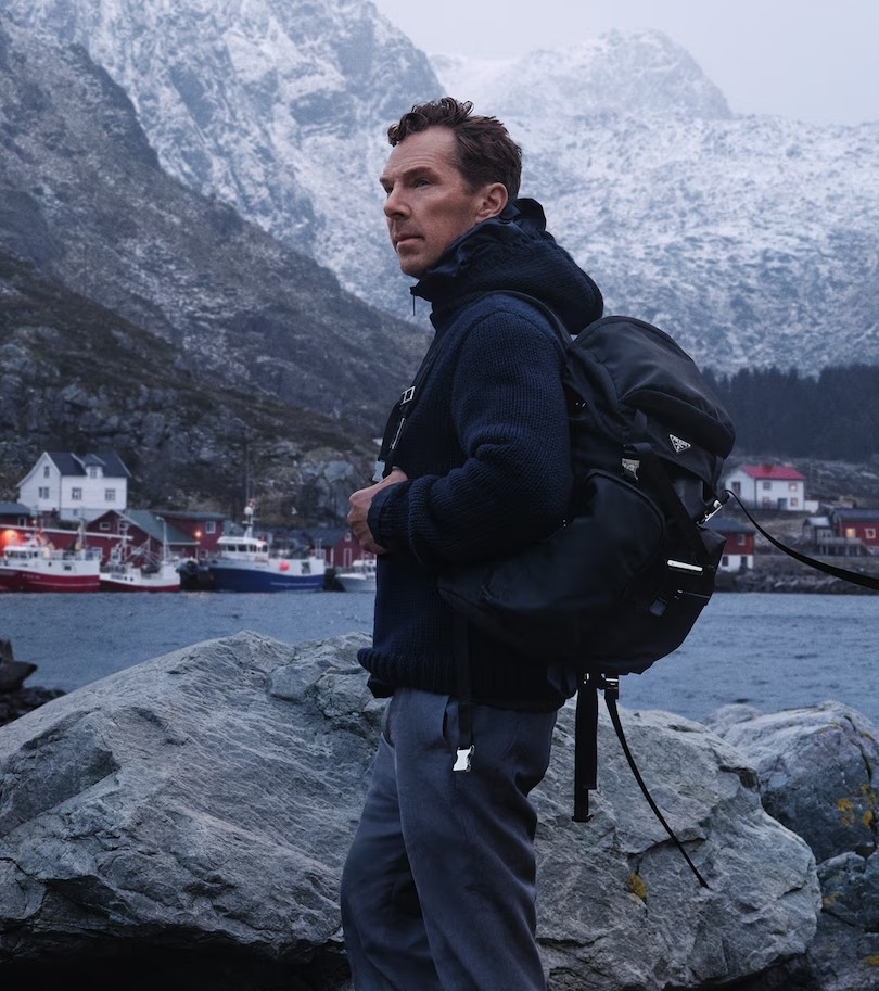 Athletic man in a sleeveless black outfit posing confidently outdoors under a bright blue sky, with a close-up of navy and salmon-colored men’s running shoes in the foreground.