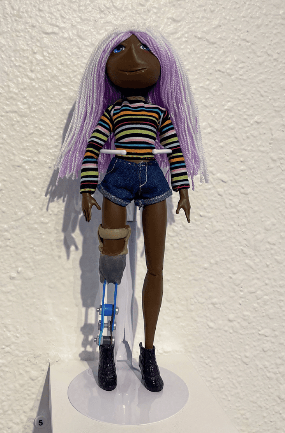 Savannah, a handcrafted doll by repaints_doll, features lavender hair, a striped shirt, denim shorts and a prosthetic leg, created for the Echoes of Resilience exhibition.