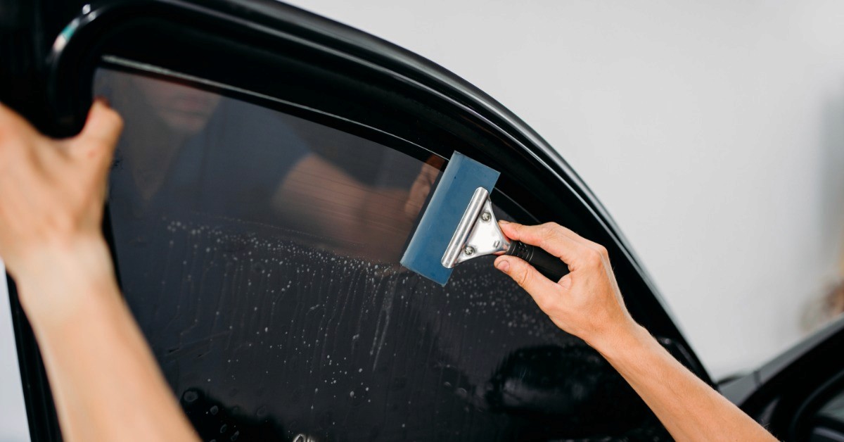 A person's hands smoothing dark tint film across a car window with a squeegee, with water droplets on the glass.