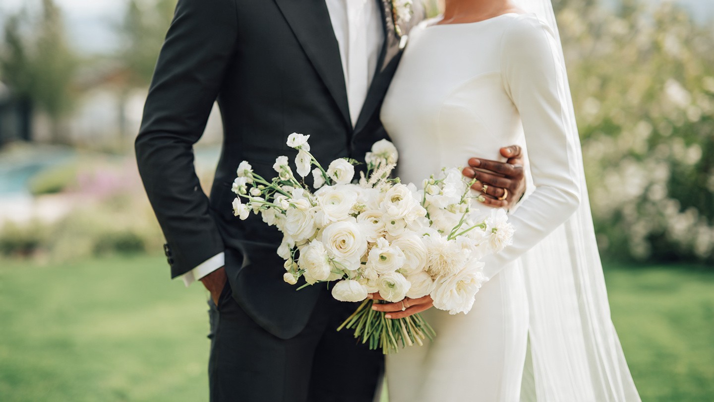 Romantic outdoor shot of an interracial couple; the groom in a black suit with hands in pockets stands beside the bride in a fitted white long-sleeve gown and veil, together cradling a full, textured bouquet of white and pale ranunculus; lush garden and pool visible in soft-focus background.
