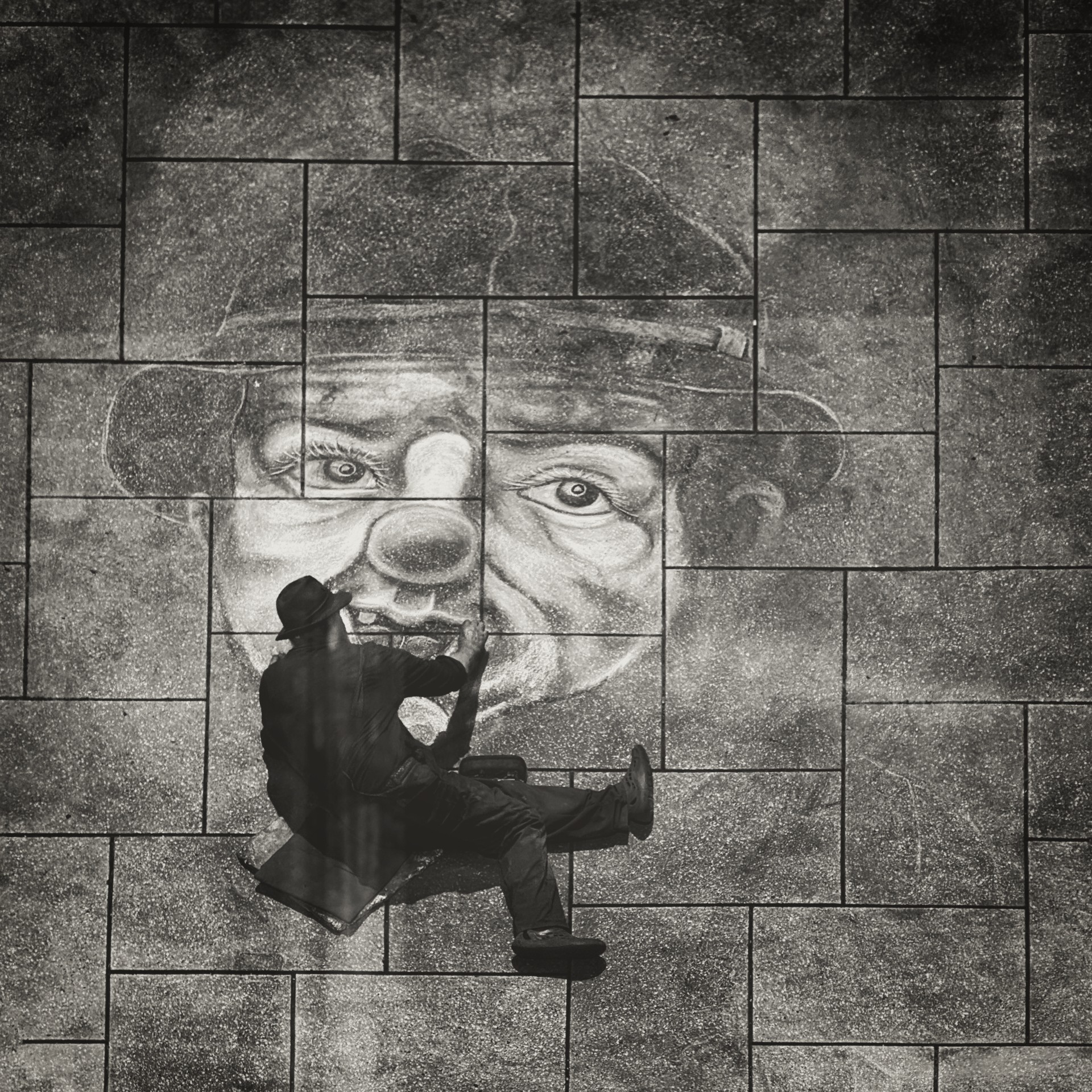 Black-and-white street art photo of a chalk artist creating a large 3D clown pavement drawing on stone tiles; ideal for content about sidewalk chalk art, urban street artists, public art installations, and photorealistic pavement murals.