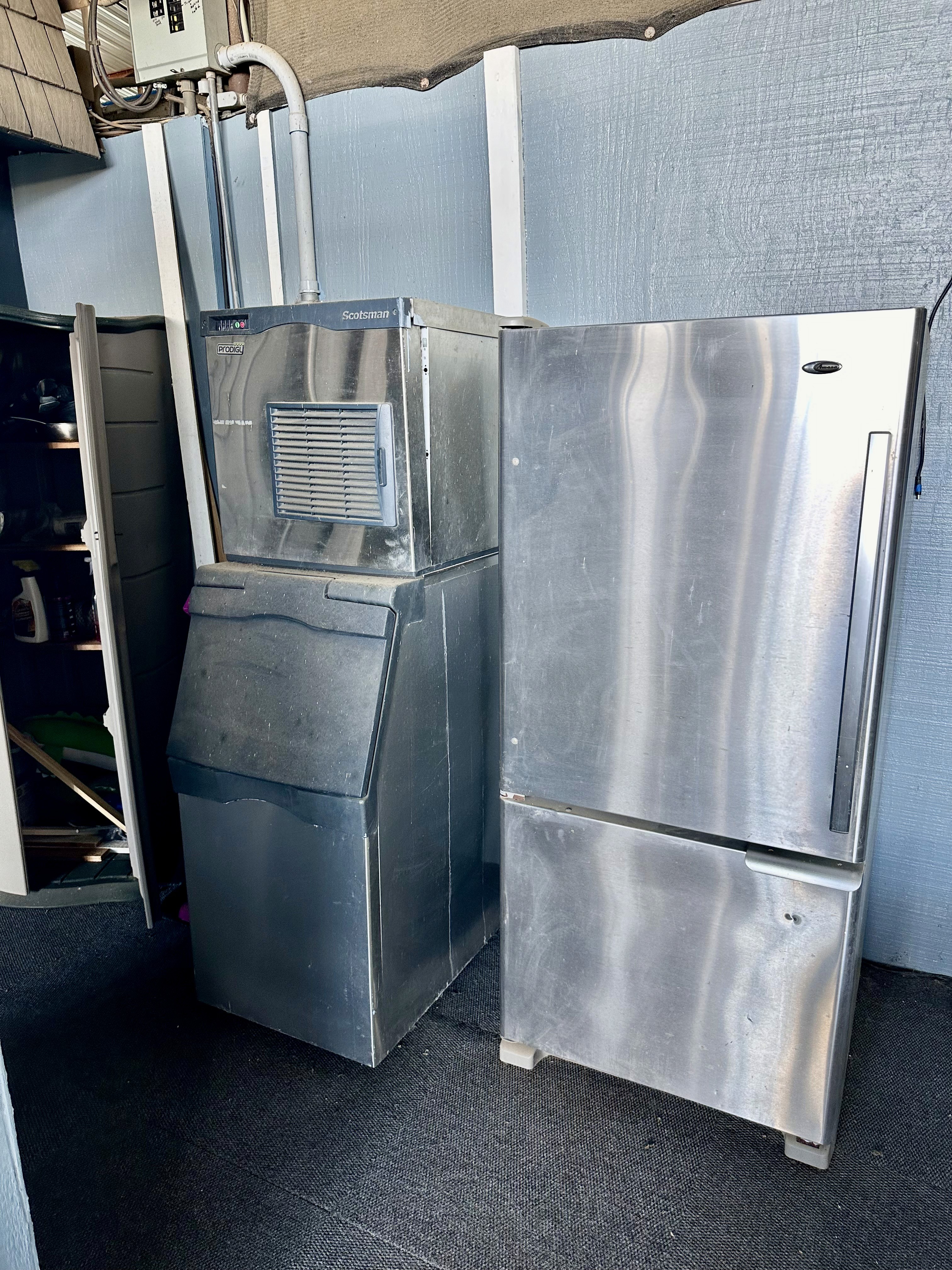 The image shows a commercial ice machine and a stainless steel refrigerator in a utility area with visible electrical connections, set against a backdrop of metallic panels, conveying an industrial setting.