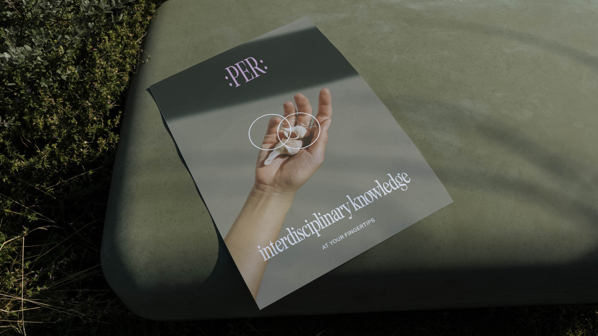 A book titled “interdisciplinary knowledge” lies open on a dark green surface. The cover features a hand holding a white circular object with concentric circles inside, and the word “PER.” appears in purple at the top left. The subtitle “AT YOUR FINGERTIPS” appears in white at the bottom right. The background includes green foliage.