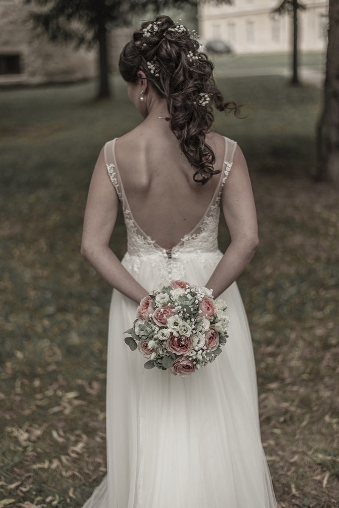 Elegant bride captured from behind holding a delicate bouquet of pastel roses — fine art wedding portrait in a serene garden. Élégante mariée photographiée de dos tenant un bouquet délicat de roses pastel — portrait de mariage artistique dans un jardin paisible.