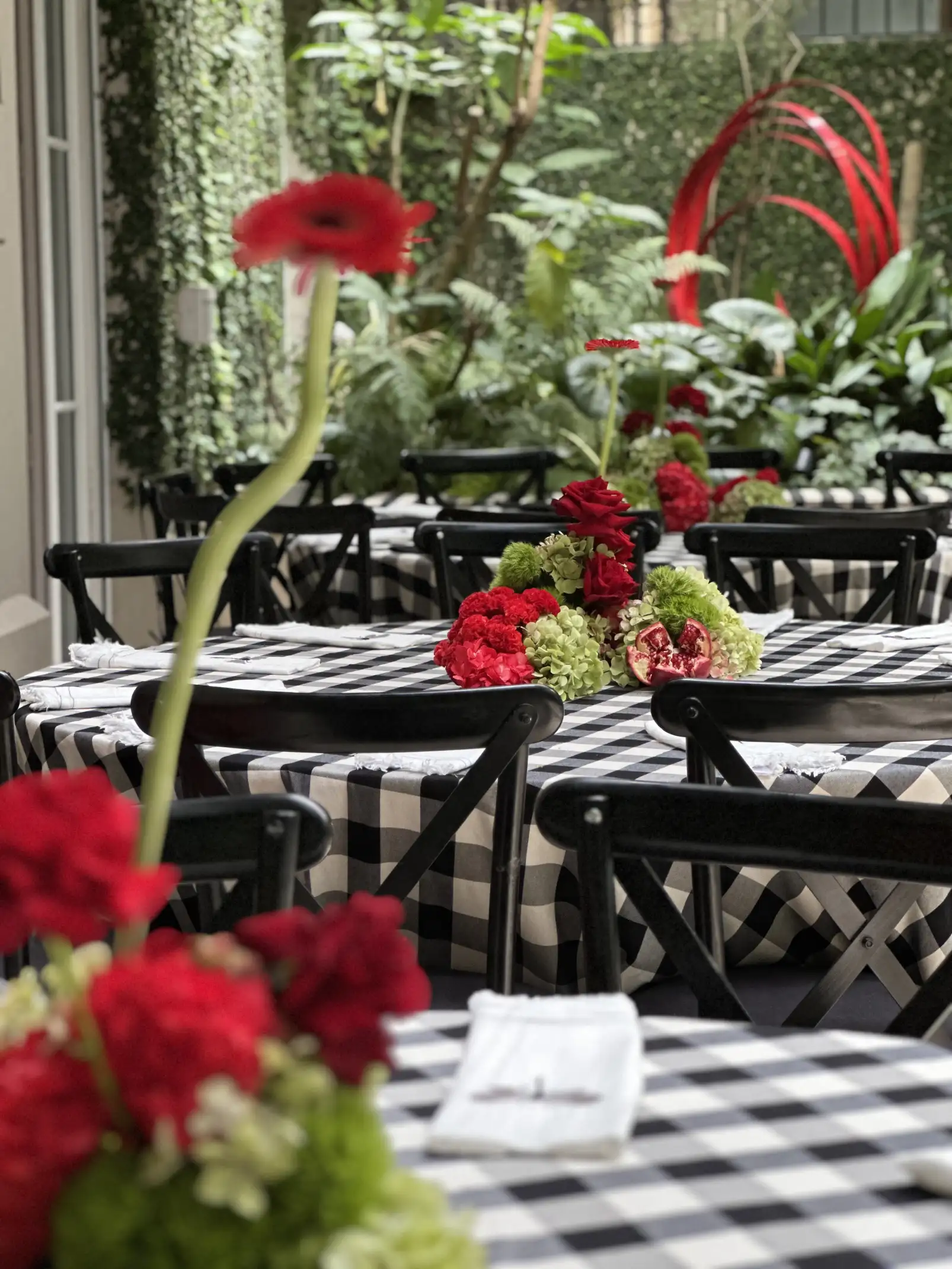 Mesas decoradas con centros florales rojos y verdes sobre manteles a cuadros en patio rodeado de vegetación para evento sofisticado.