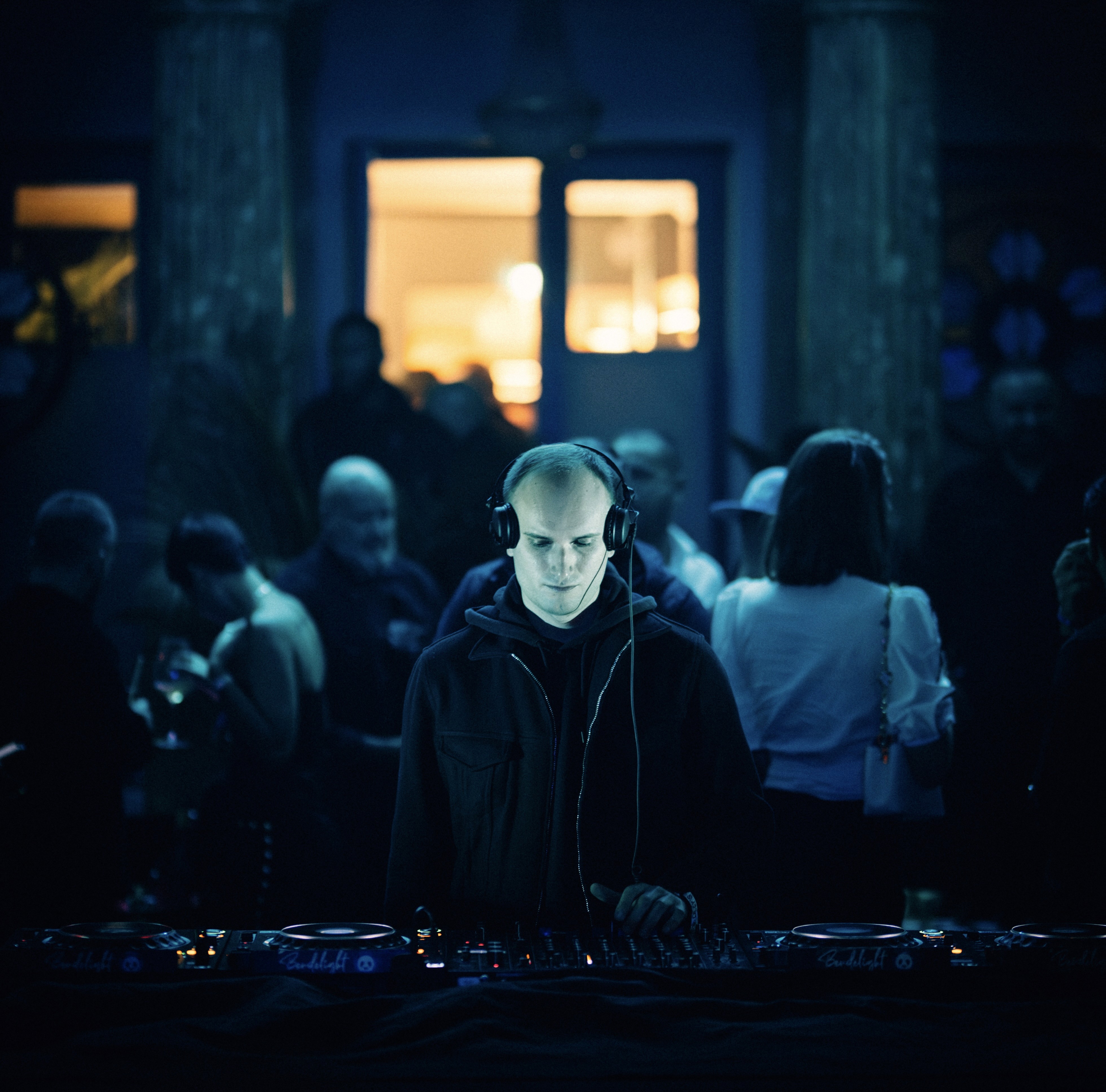 Photographie de l'artiste Jarod portant un casque audio autour du cou, debout au centre d'une foule floue dans une ambiance de club nocturne aux tons sombres et bleutés.