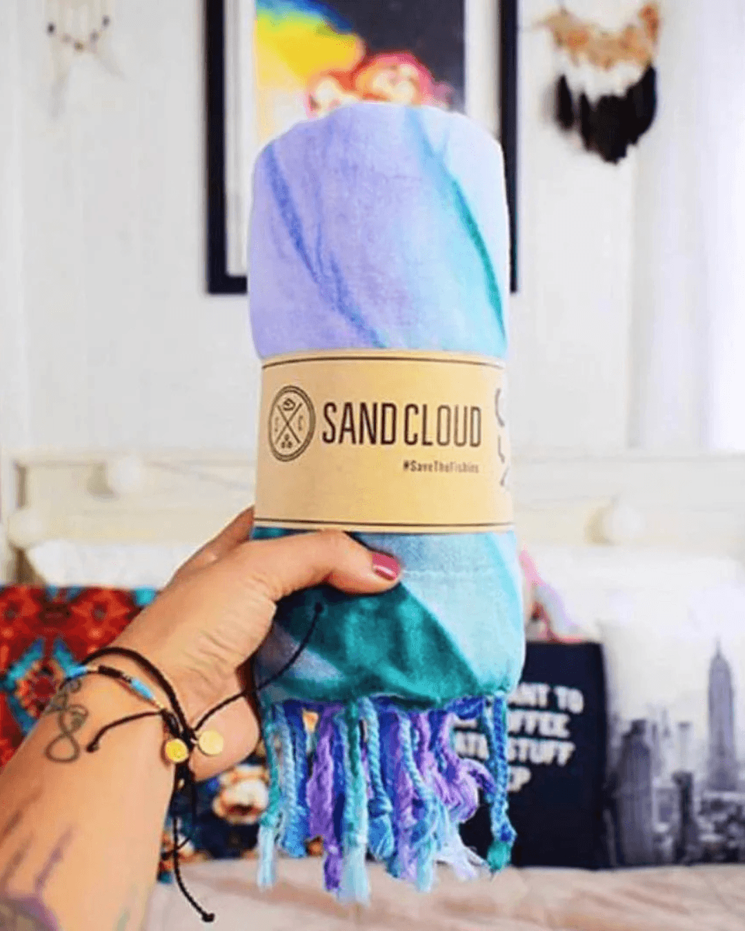 A hand holds a rolled towel in pastel colors, with a label, against a blurred background of decor and furnishings.
