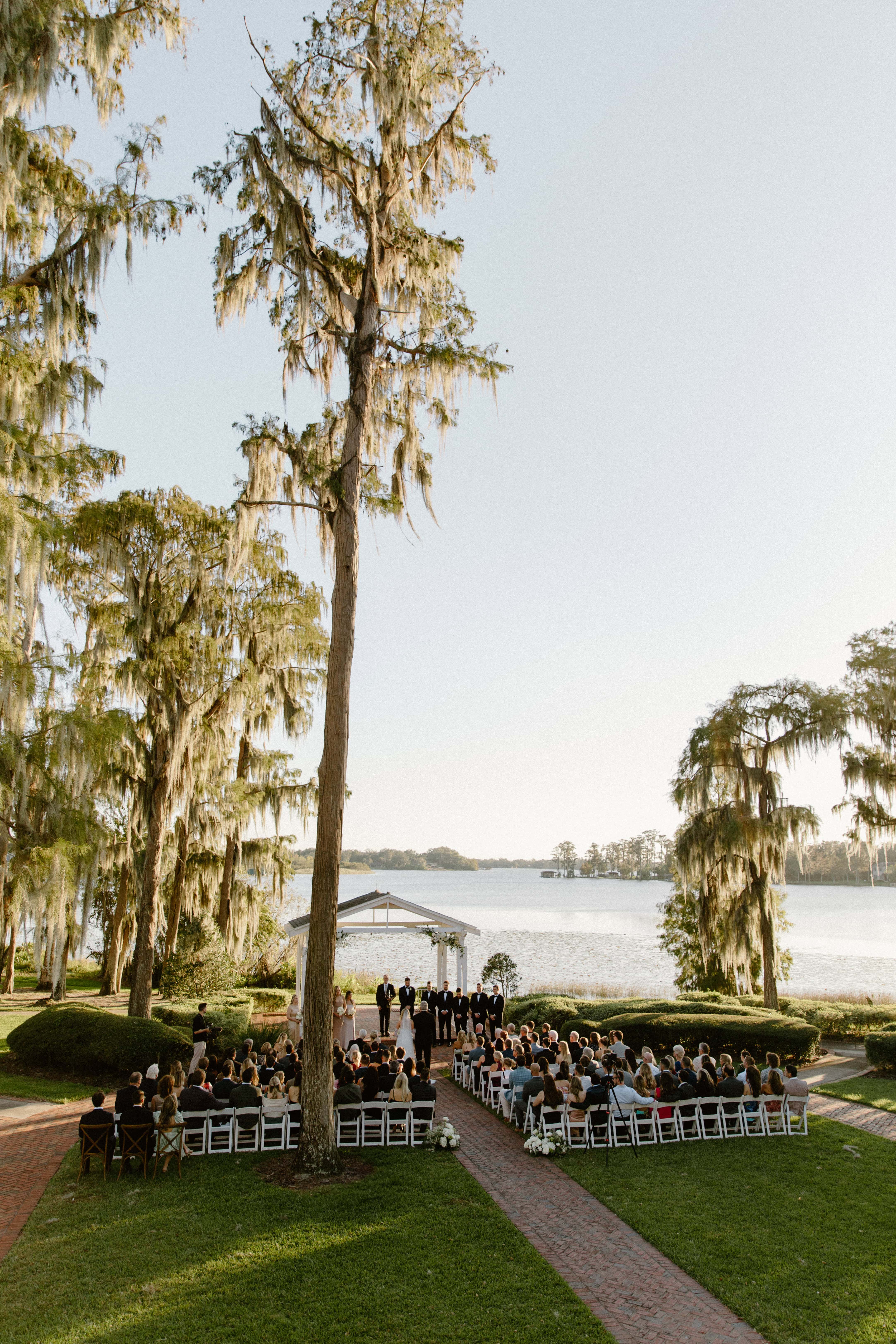 A photo of an outdoor wedding ceremony with chairs set up on a grassy lawn facing a body of water in the distance. The sun is low in the sky.