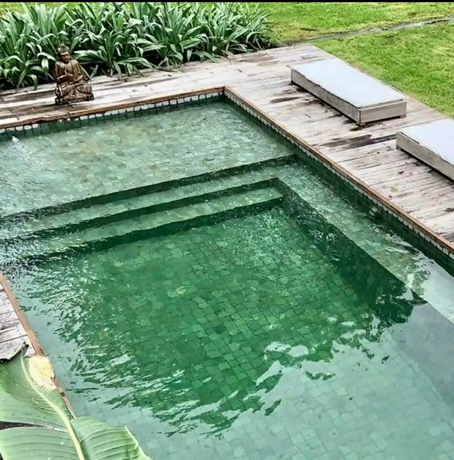 Piscina de lujo revestida con auténtica Piedra Bali verde esmeralda, acompañada de un deck de madera natural y decoración zen en el exterior.