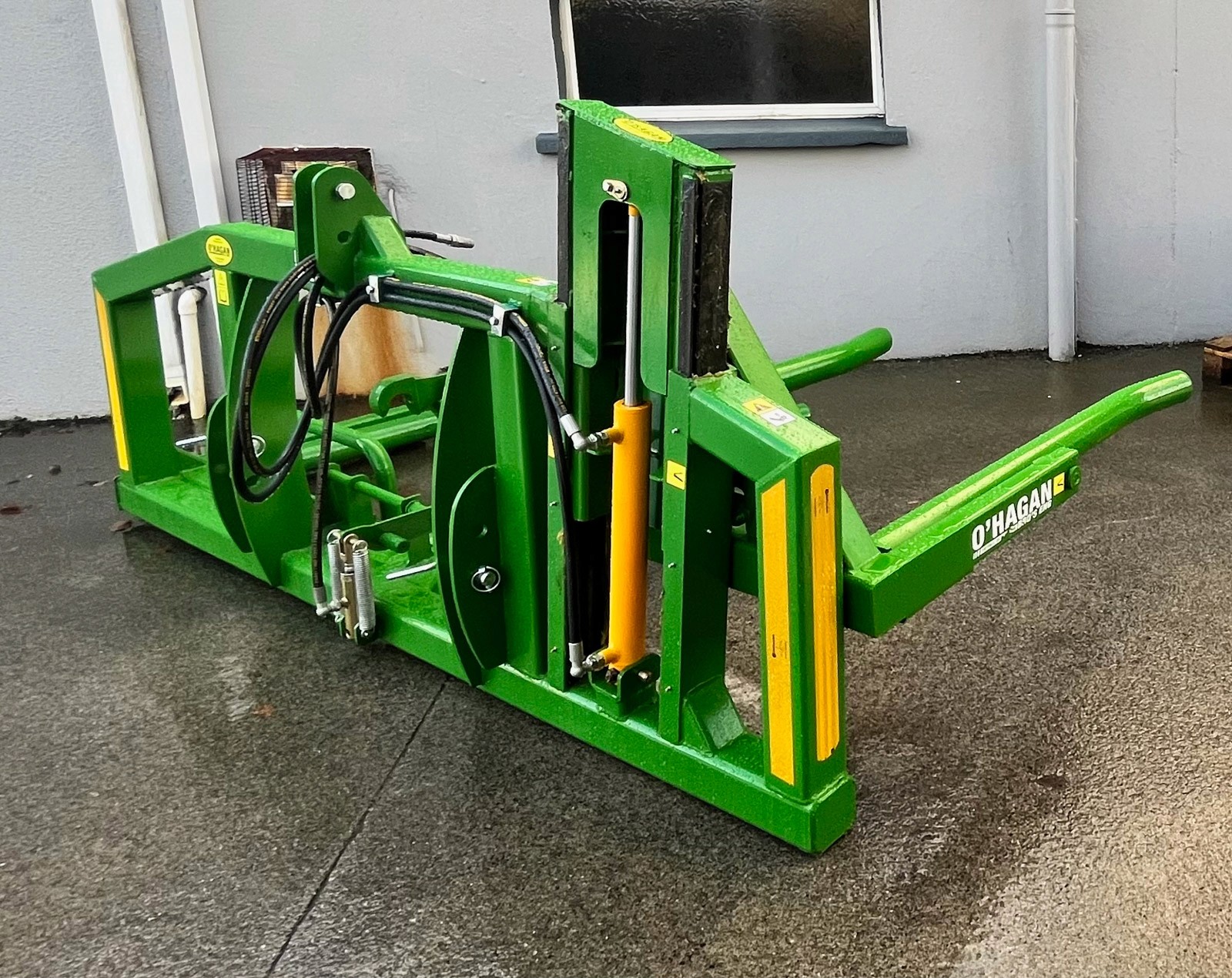 Green hydraulic Bale handler outdoors on a concrete floor beside a building with a window