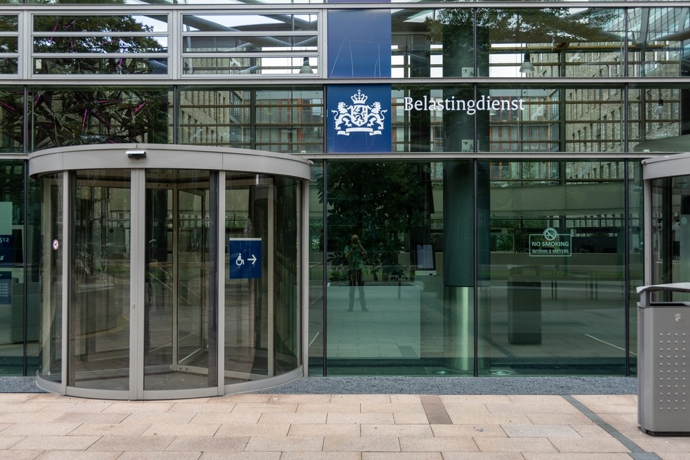 Ingang van een overheidsgebouw met het logo van de Rijksoverheid en een draaideur, weerspiegeling van de omgeving zichtbaar in het glas