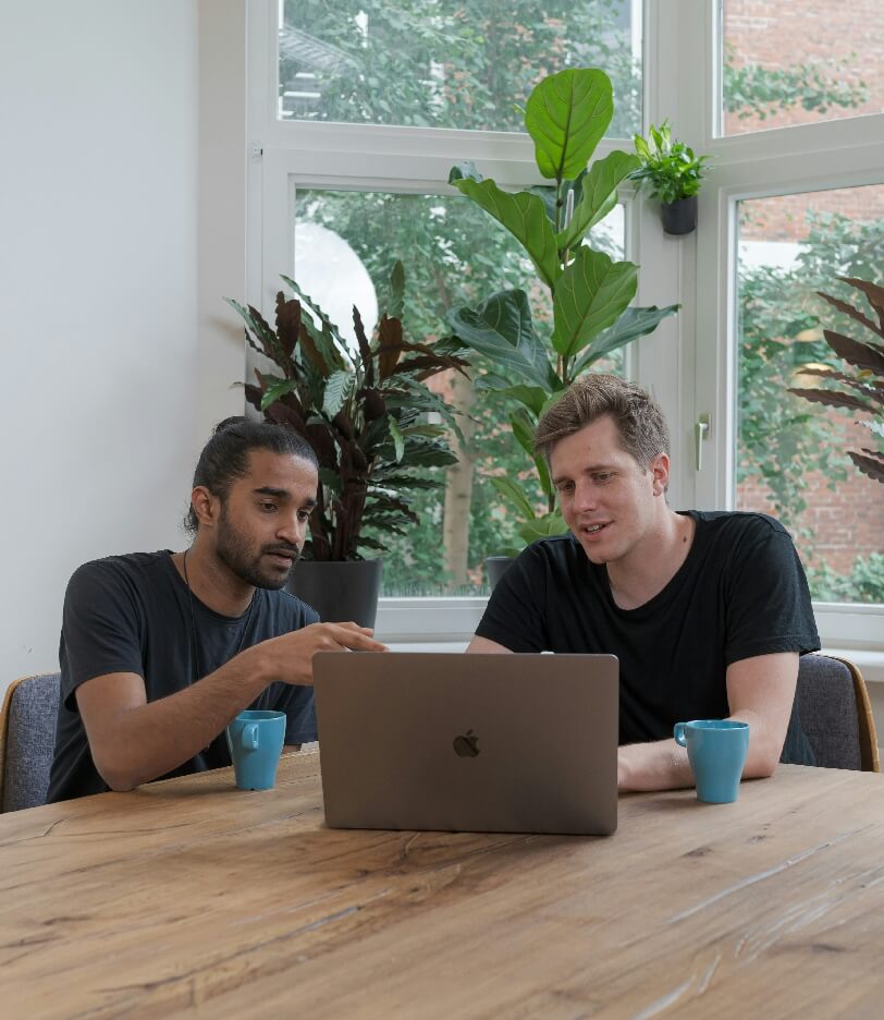 Two colleagues discussing work at a table with laptops in a bright office space.