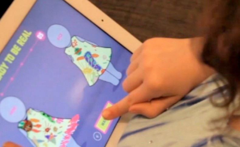 A child's hand points at a colorful, cartoon-style dress displayed on a tablet screen. The dress has various patterns and shapes, including what appears to be a cat or similar animal design. The screen shows a purple background with the text "BABY TO BE REAL" at the top left and a green button at the bottom center. The child is wearing a light blue and white striped shirt. The tablet is resting on a dark surface.