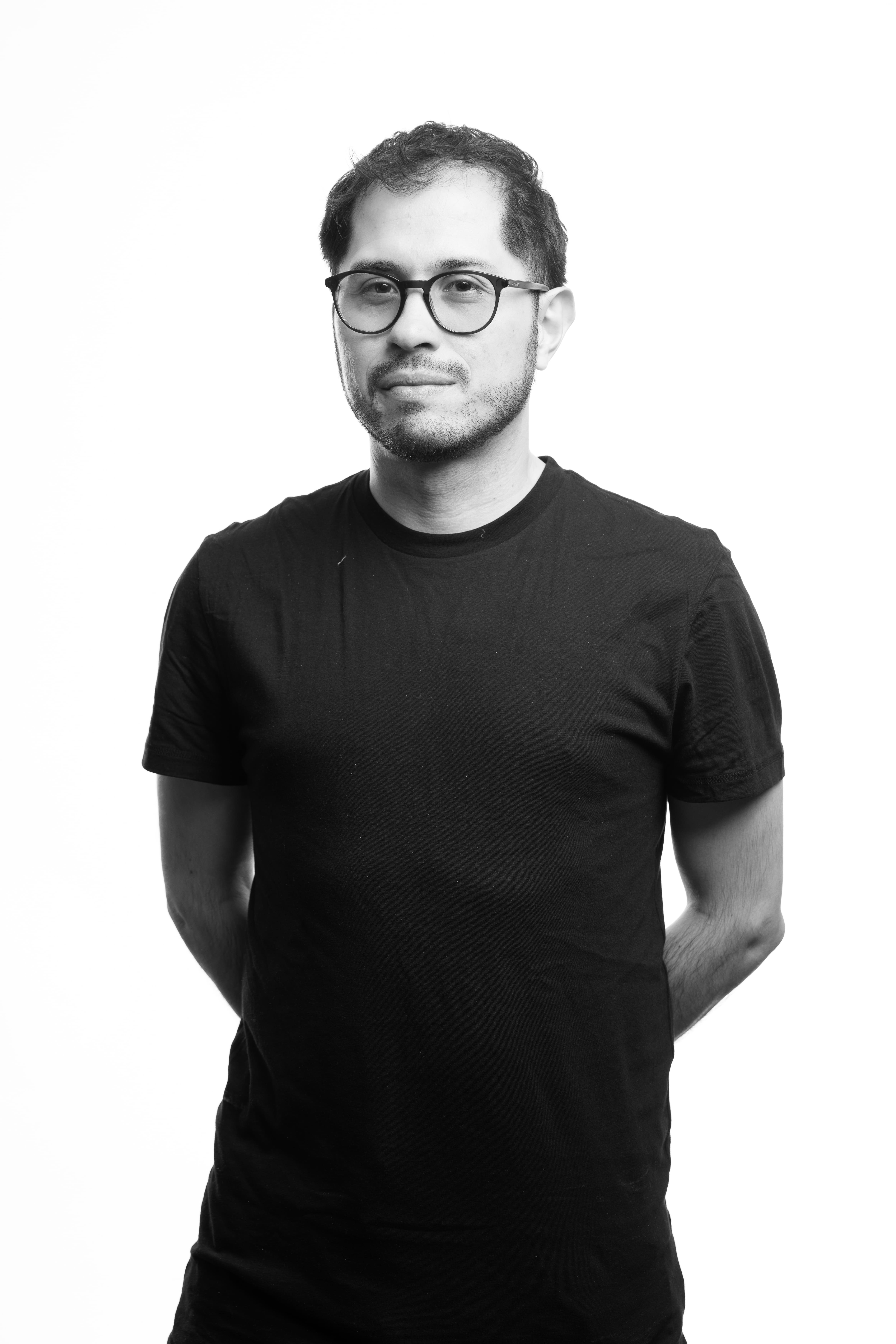 A man with glasses and a short beard stands casually with hands behind his back, wearing a black t-shirt, against a plain white background.