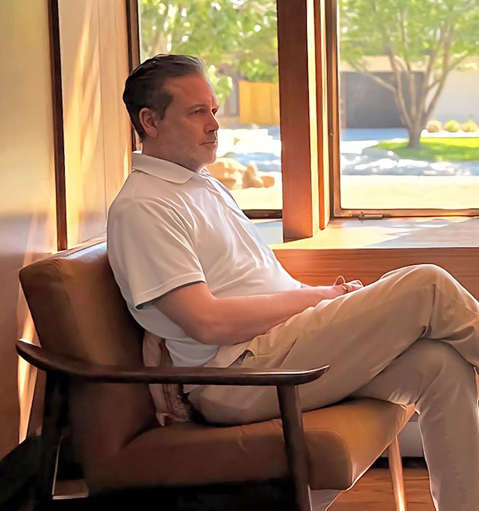 Abe Jasinowski (Abe Zemi) sitting in a wooden chair near floor-to-ceiling curtains and a large window overlooking a garden.