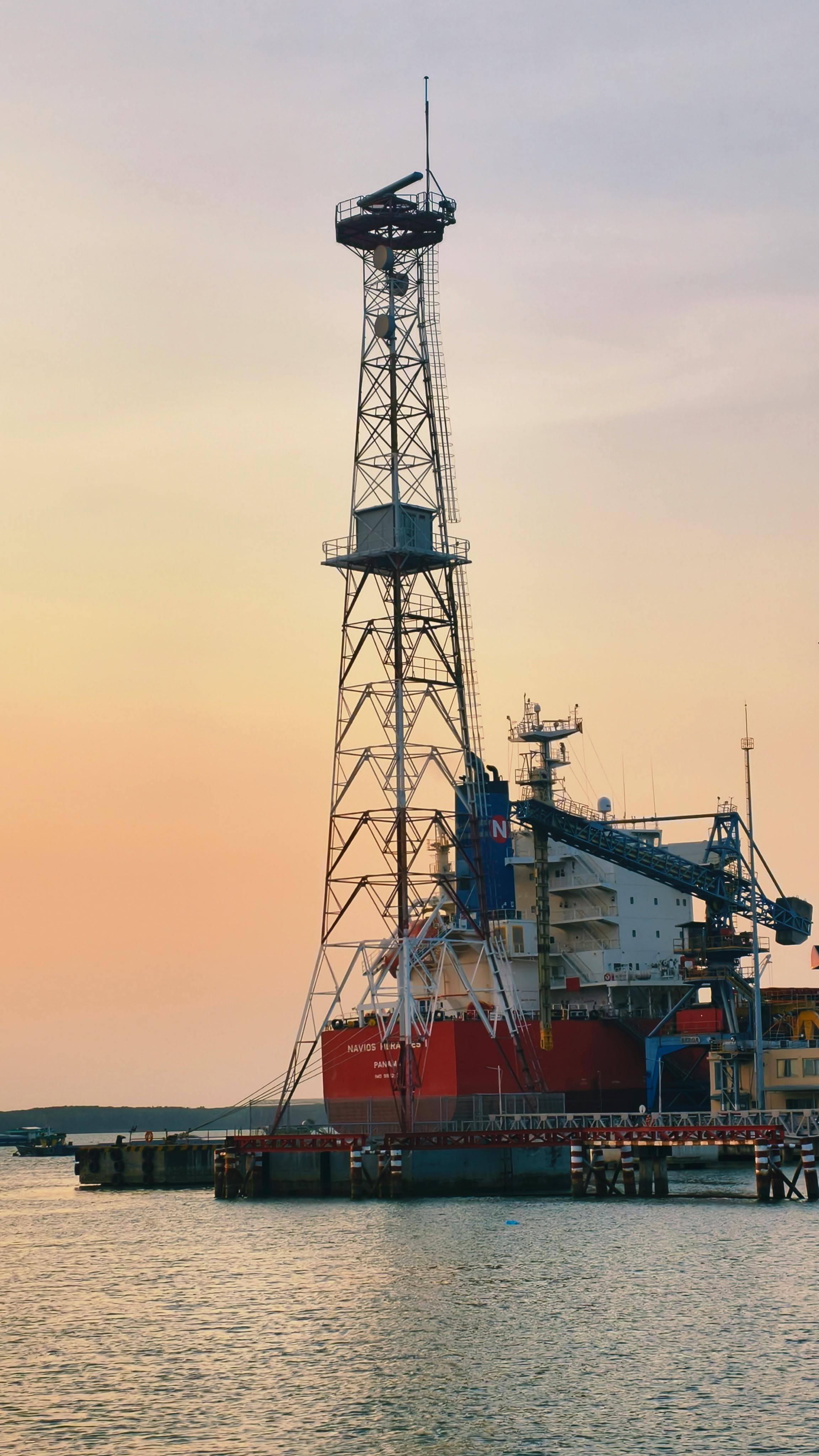 A tall offshore drillship with a prominent derrick tower docked at a marine port during a pale sunset.