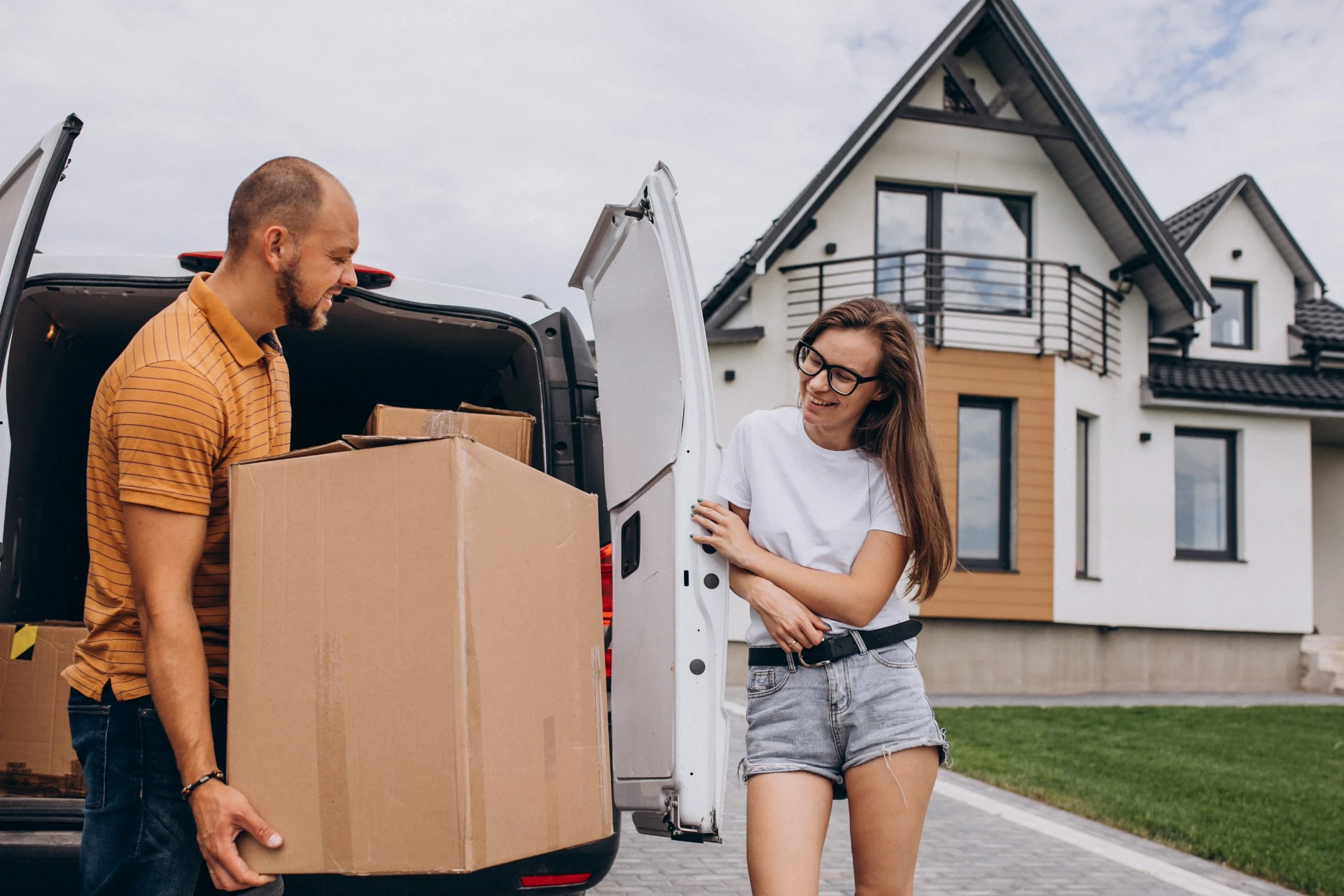 A man in a yellow striped shirt unloads a cardboard box from a white delivery van while a smiling woman in glasses watches outside their new modern home.